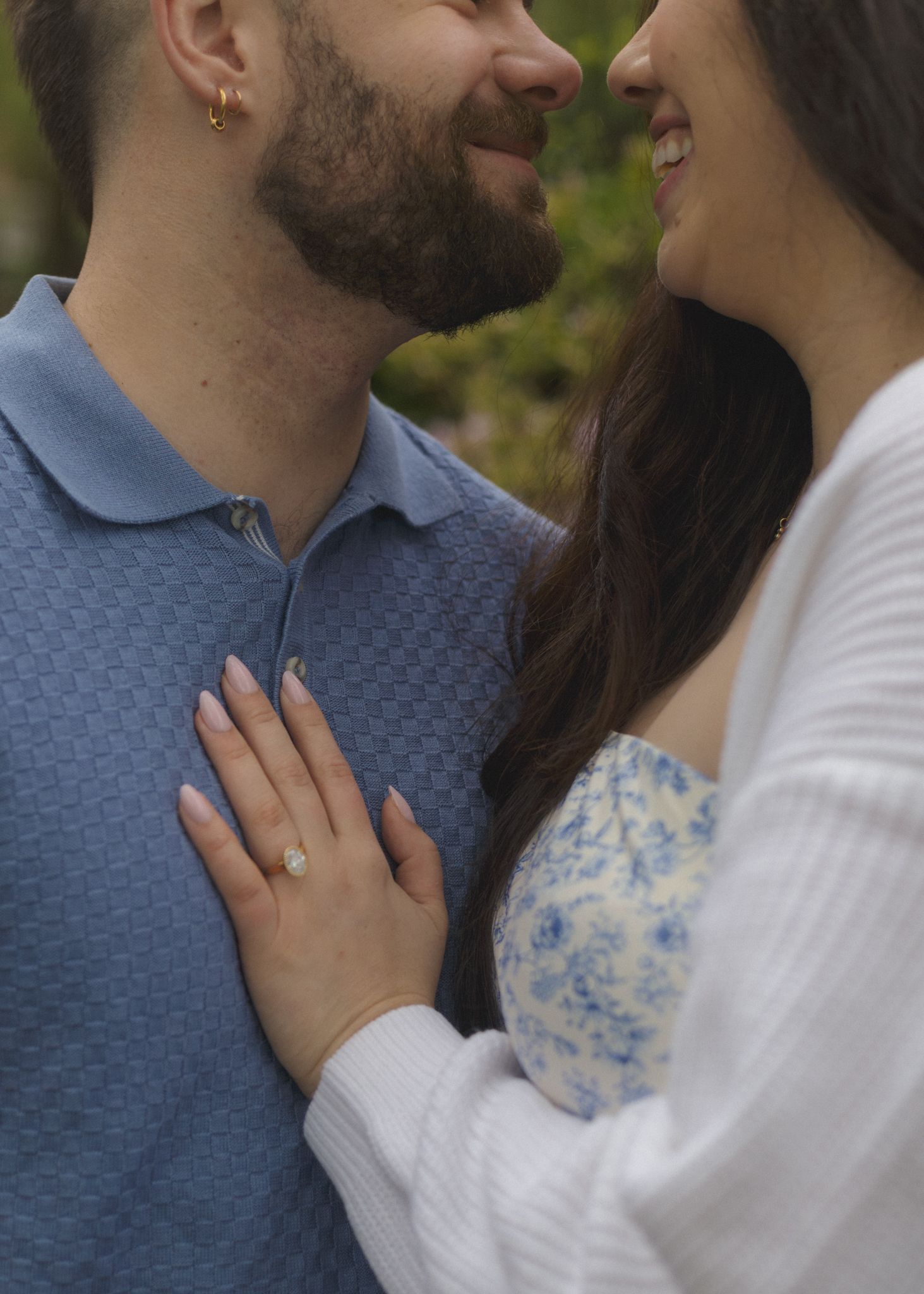 A smiling couple shares a romantic moment, the woman wearing an engagement ring resting her hand on the man's chest.