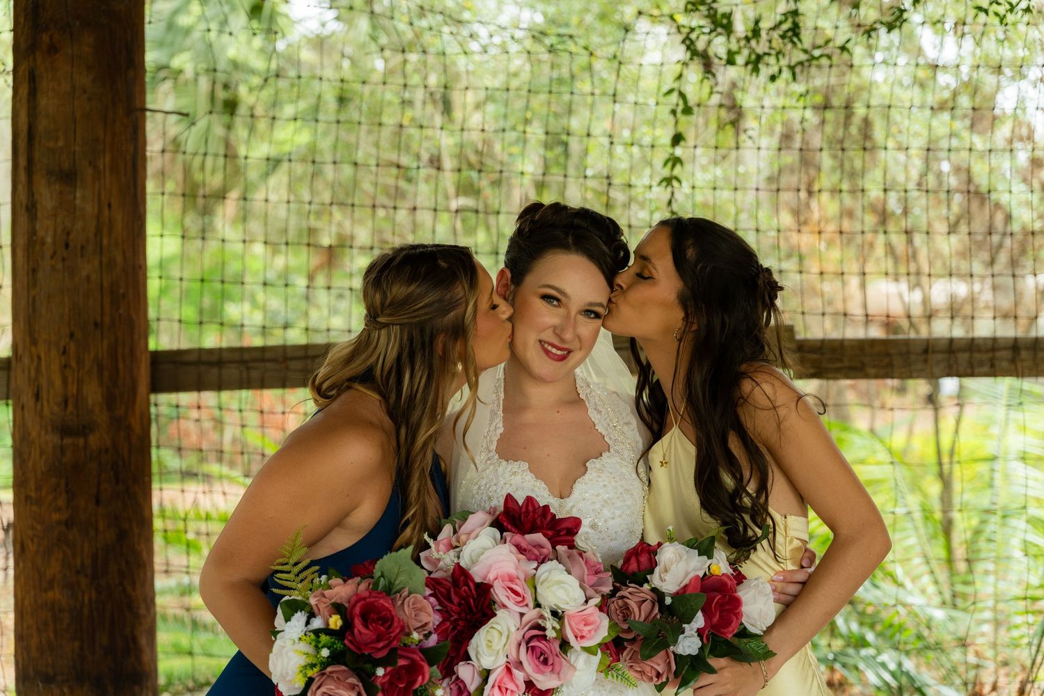 Bride in white lace gown holds pink and red bouquet while two bridesmaids in blue dresses kiss her cheeks.
