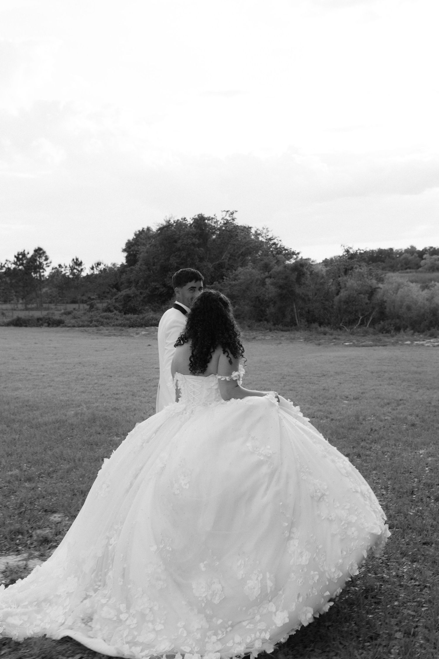 Bride in floral ball gown and groom in white tuxedo embrace in black and white field photo.