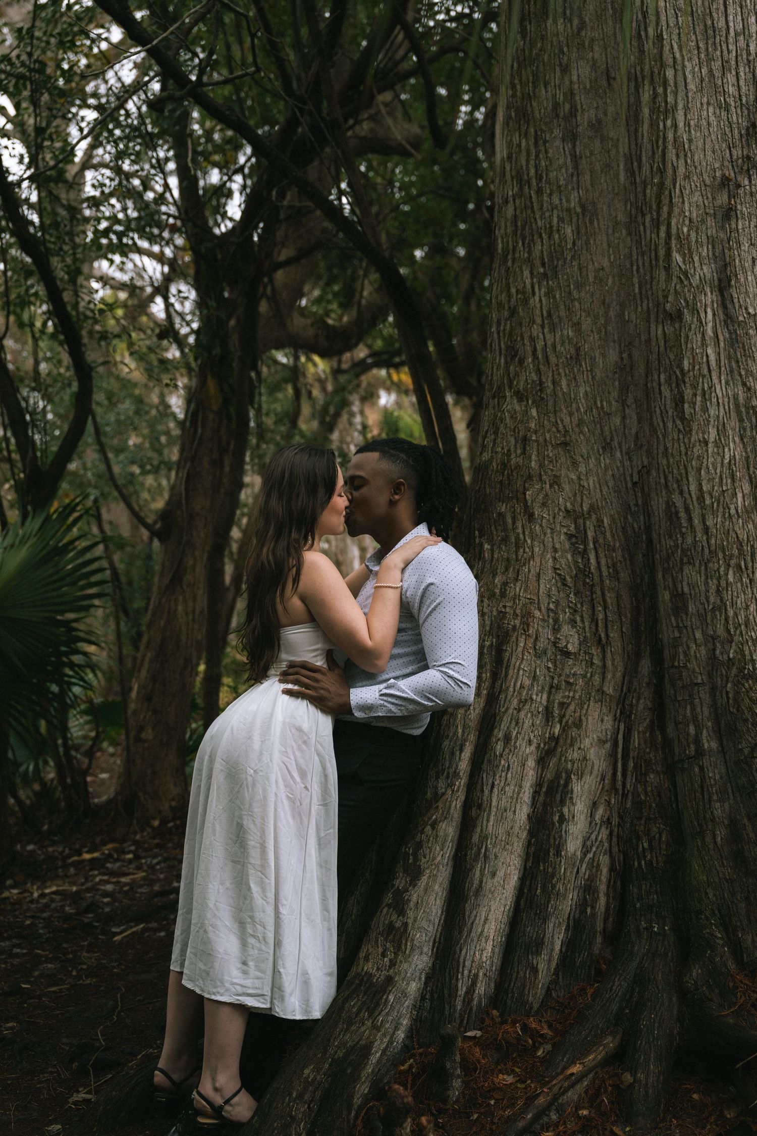 A romantic couple embraces against a large tree trunk in a lush forest setting with filtered sunlight.