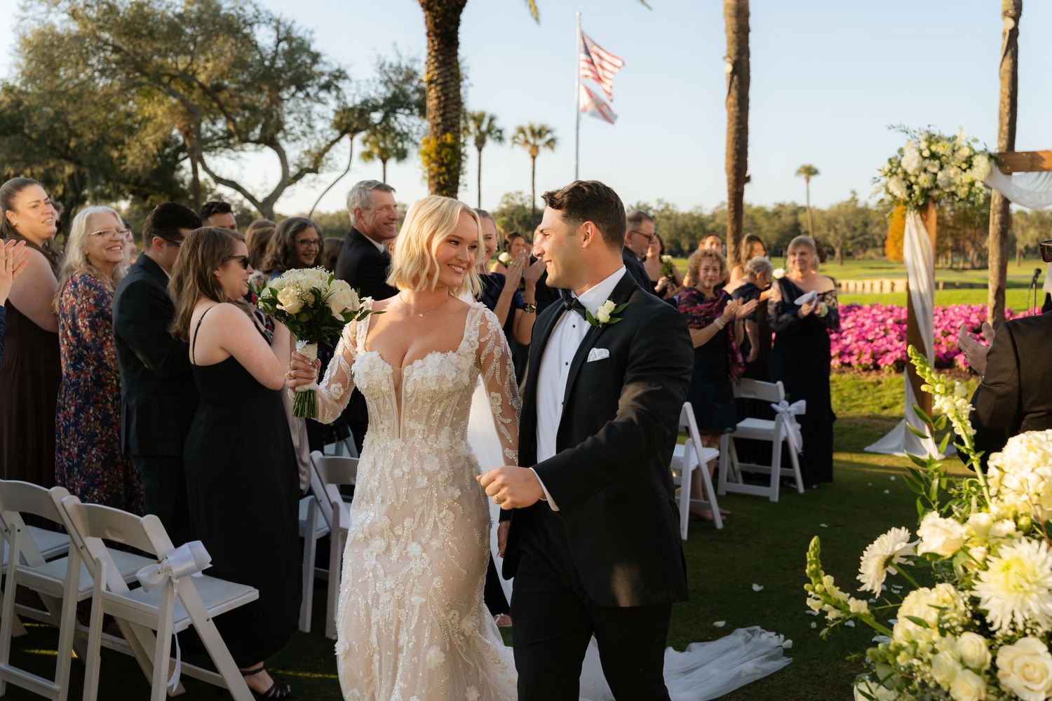 Smiling newlywed couple walks back down aisle after outdoor wedding ceremony with cheering guests.