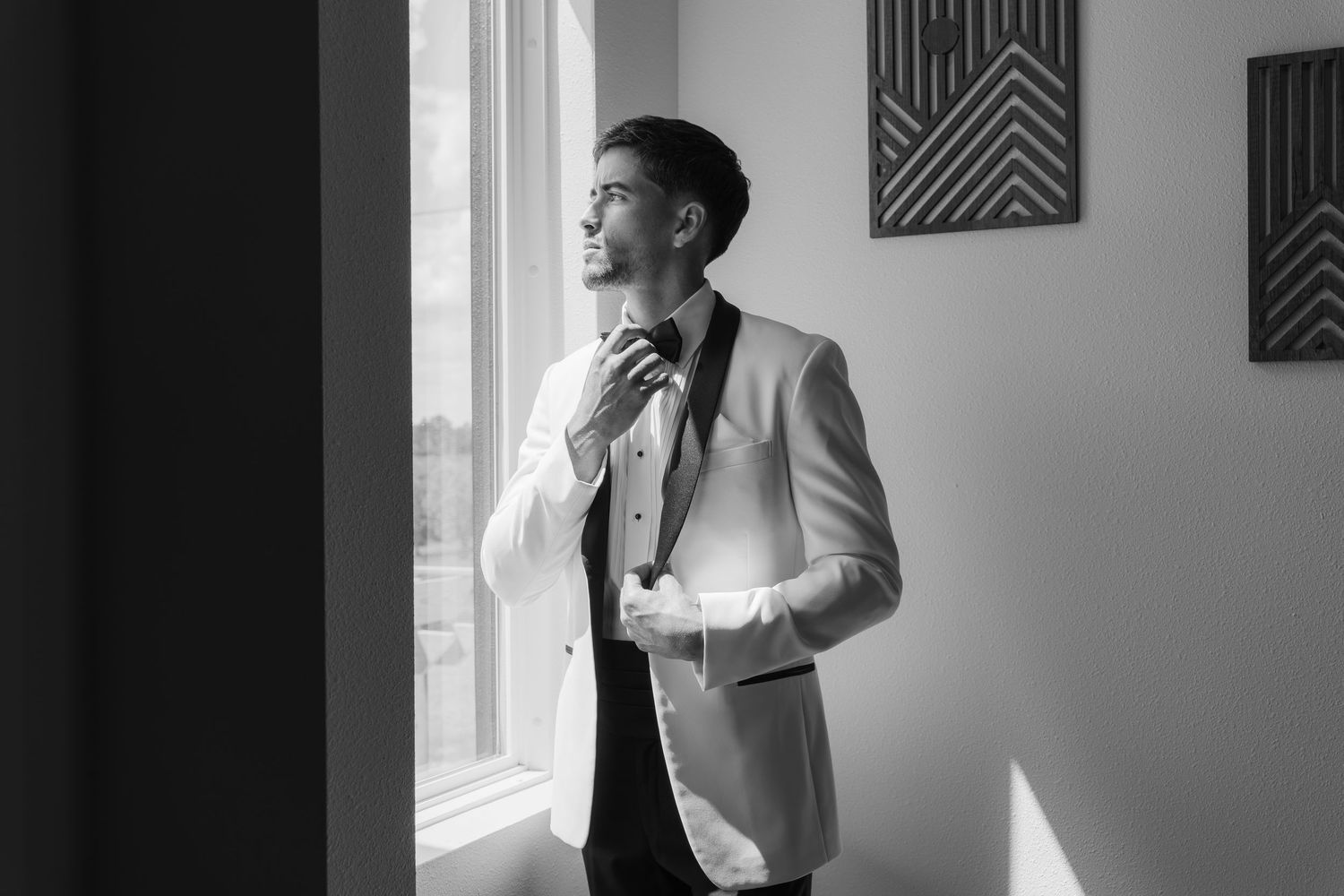 Black and white photo of a groom in a white tuxedo adjusting his bow tie by a sunlit window.