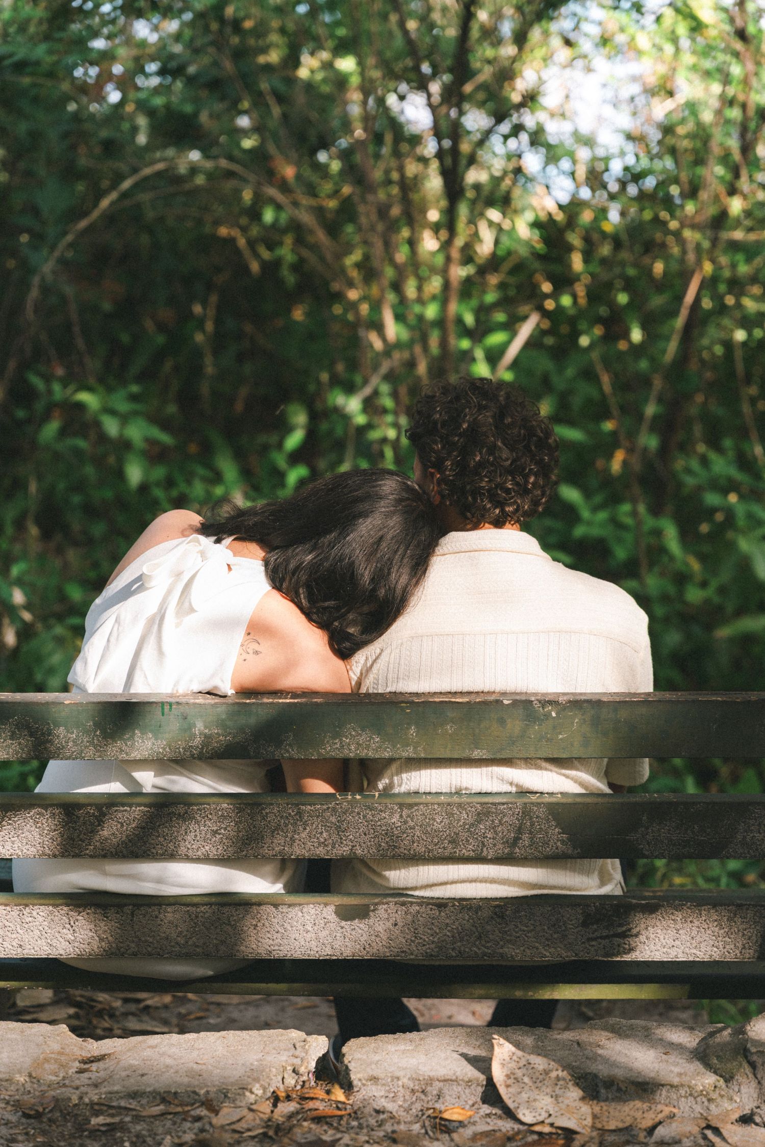 Two people rest together on a wooden bench surrounded by lush greenery in a peaceful outdoor setting.