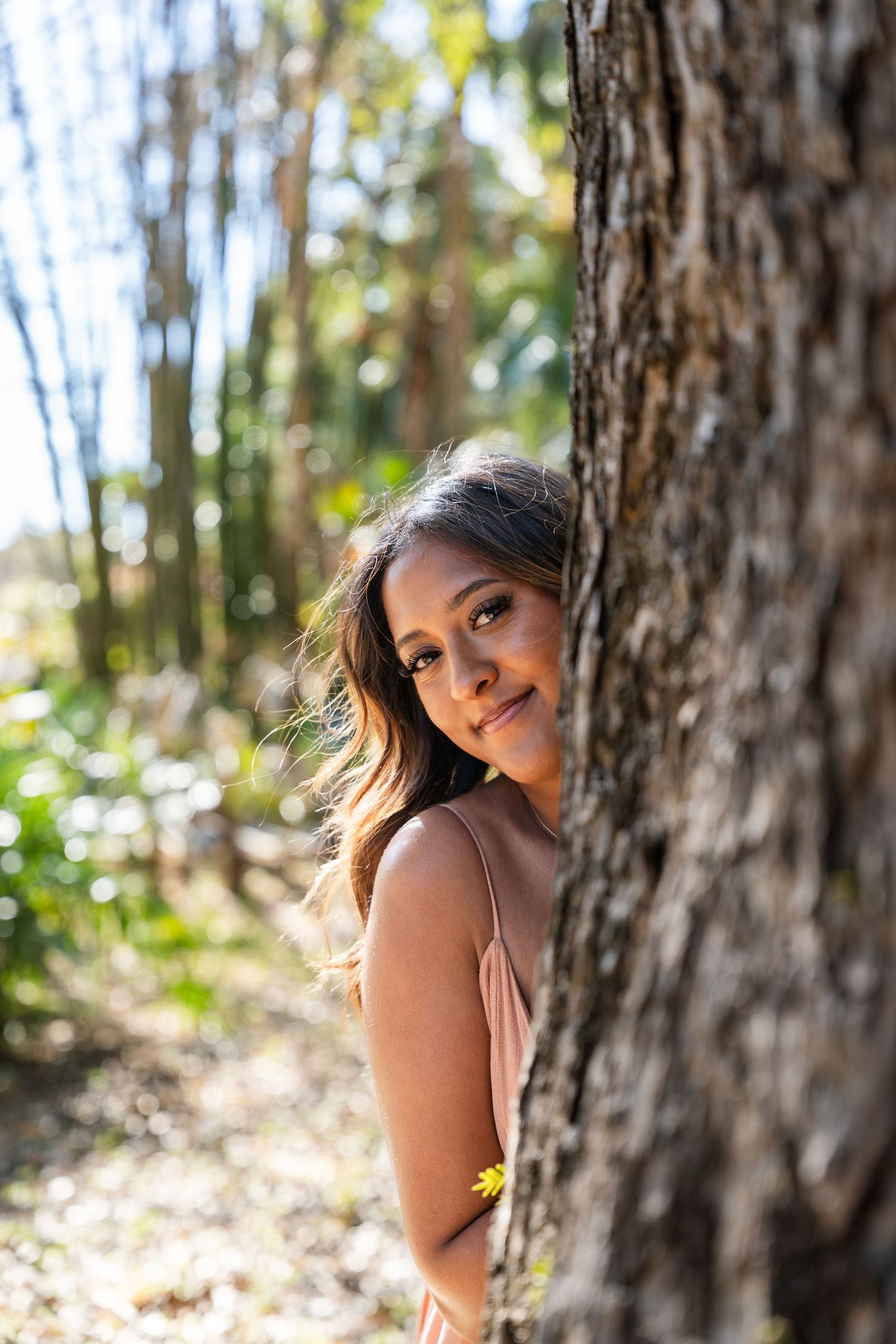 Young woman with long hair peeking from behind a tree trunk in a sunlit forest, smiling softly.