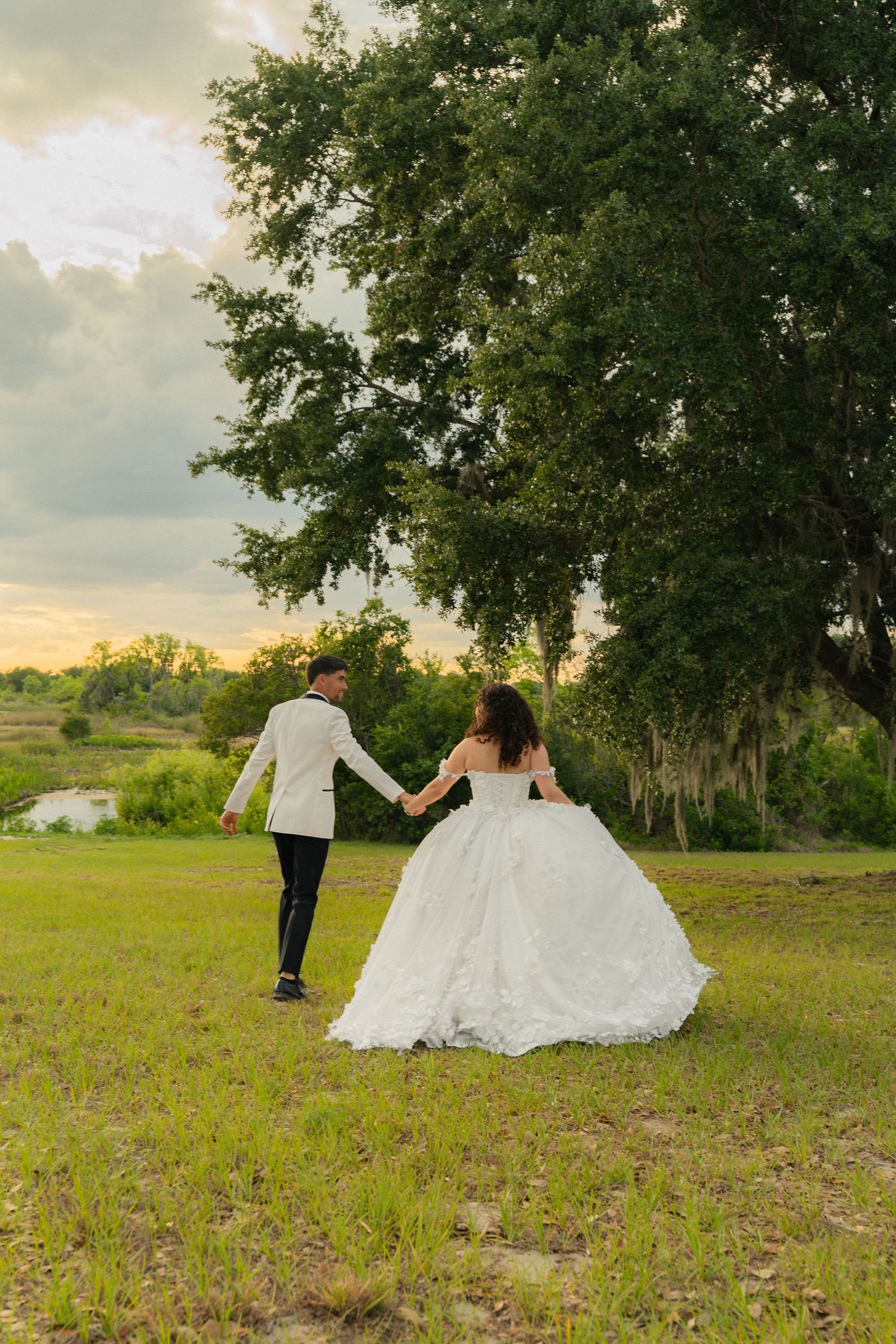 Bride and groom walk hand in hand across green meadow toward large oak tree at dusk.