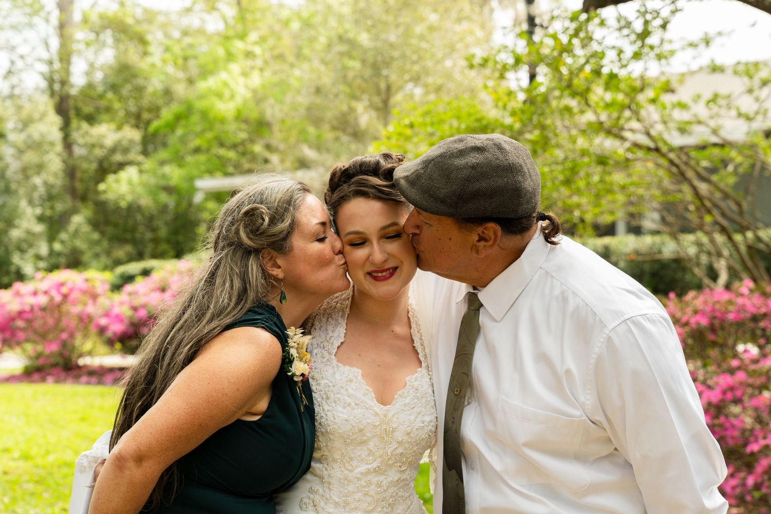 Bride receives kisses on cheeks from both parents during outdoor wedding celebration surrounded by blooming flowers.
