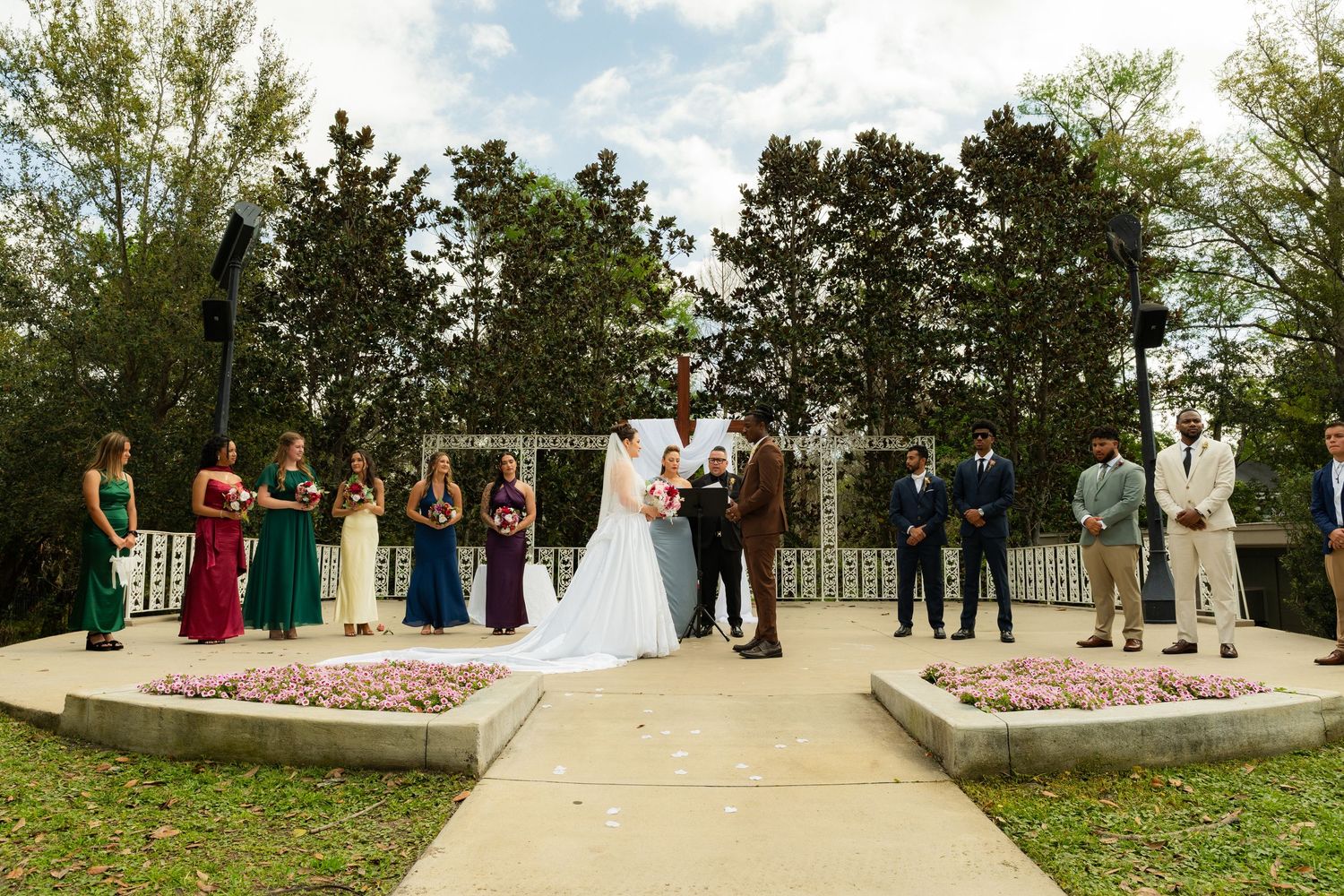 Outdoor wedding ceremony with bride and groom at altar, bridesmaids in colorful gowns and groomsmen in suits, garden venue.