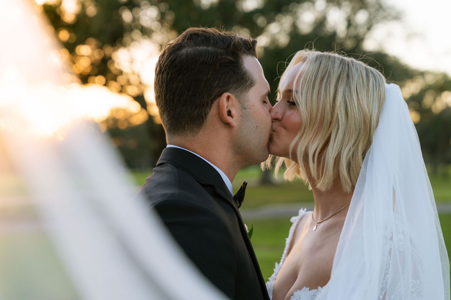 Bride and groom share a romantic kiss at golden hour, bride wearing lace gown and veil outdoors.