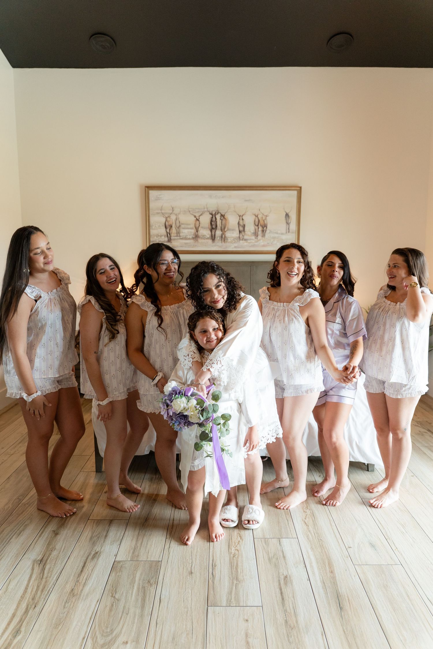 Bride in white robe holding bouquet poses with bridesmaids and flower girl in matching pajamas indoors.