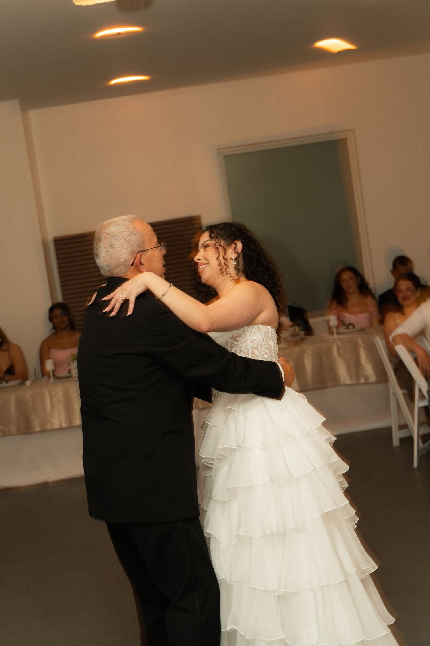 Bride in white ruffled gown shares father-daughter dance at wedding reception with guests seated in background.