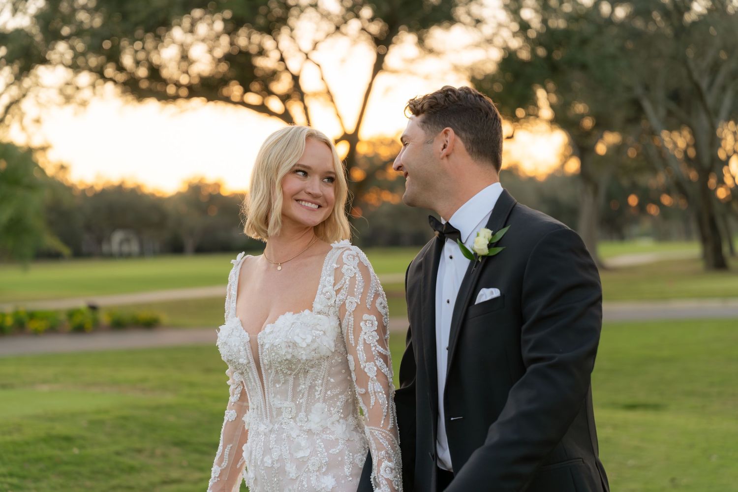 Smiling bride in lace gown and groom in tuxedo share a romantic moment at sunset during outdoor wedding portraits.