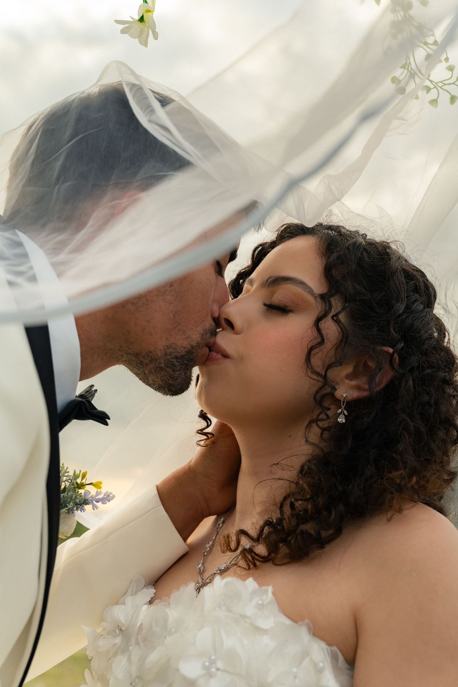 Bride and groom sharing a romantic kiss beneath a flowing white wedding veil outdoors.