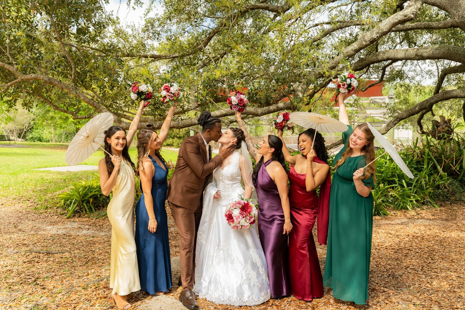 Newlyweds kiss surrounded by bridesmaids in colorful satin gowns holding bouquets under a large oak tree outdoors.