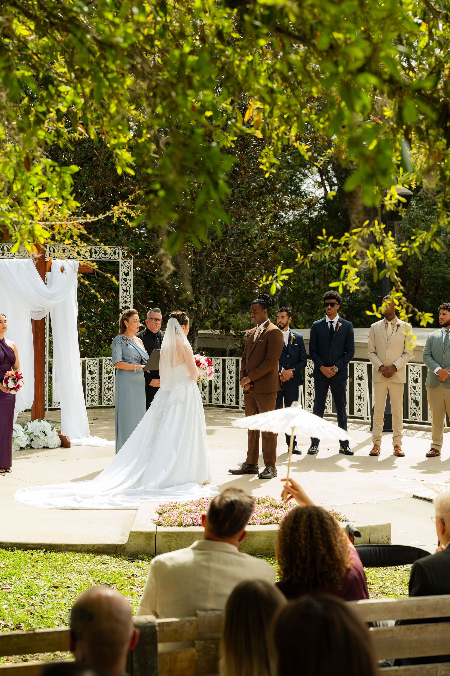 Bride in white gown exchanges vows with groom at outdoor wedding altar beneath lush green trees with seated guests.