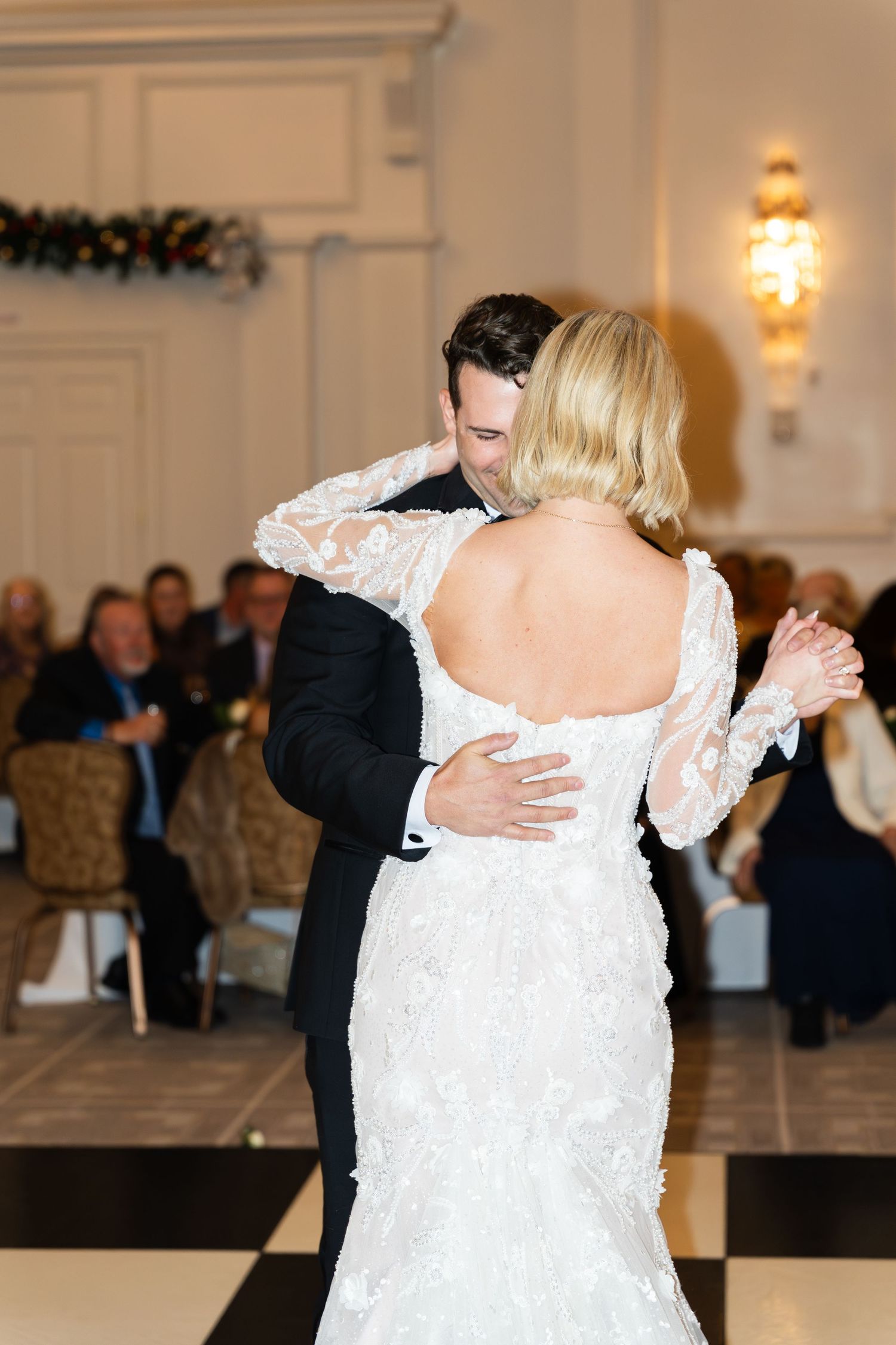 Groom in black tuxedo embraces bride in lace backless gown during romantic first dance at winter wedding reception.