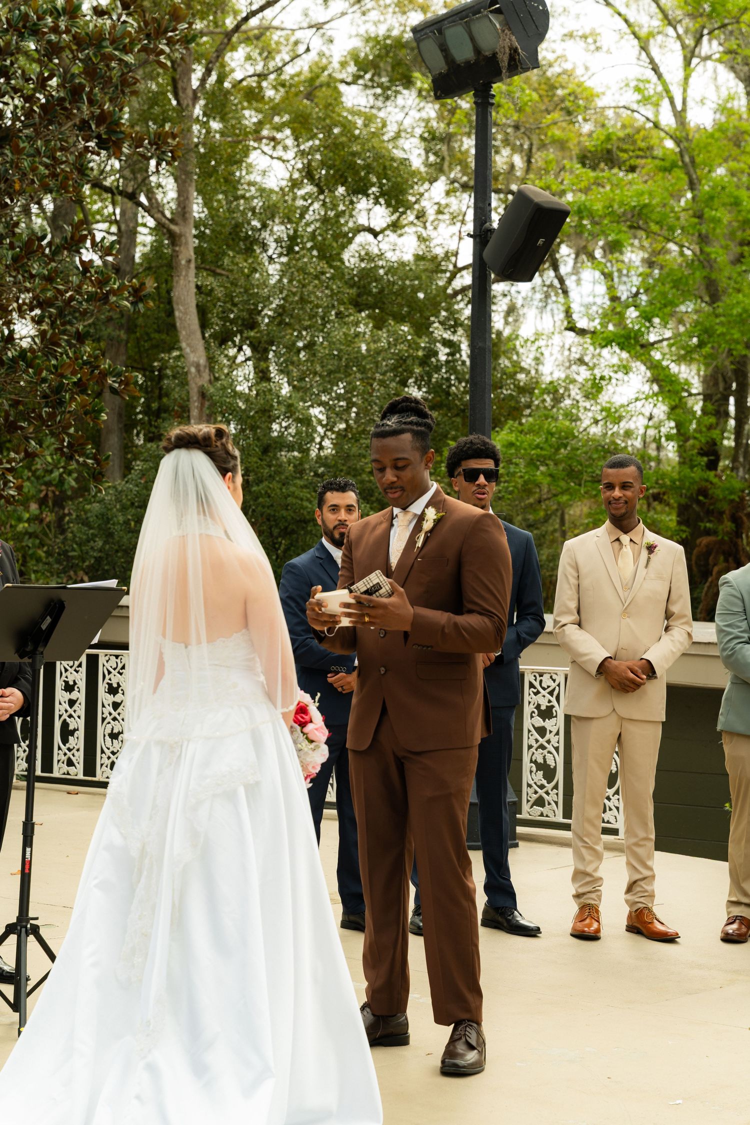 Groom in brown suit reads wedding vows to bride in white gown during outdoor ceremony with groomsmen standing behind.