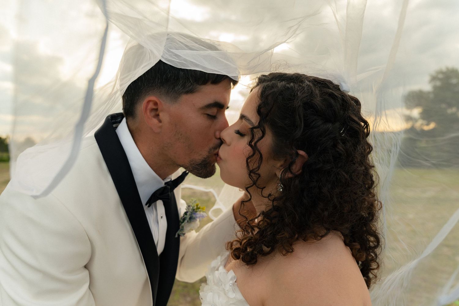 Bride and groom sharing a romantic kiss under a flowing white veil at their outdoor wedding ceremony.