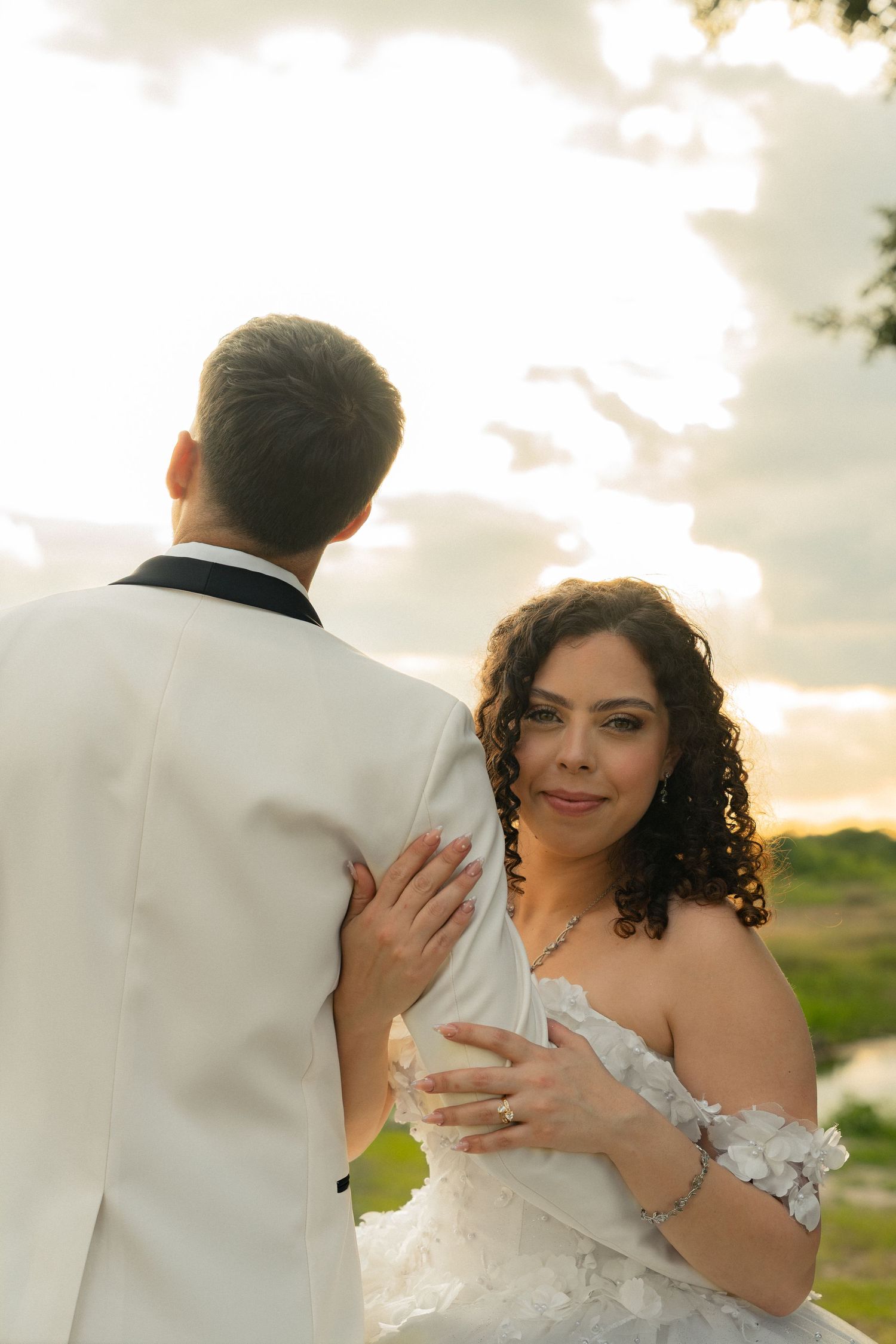 Smiling bride in off-shoulder floral gown embraces groom during golden hour wedding portraits.