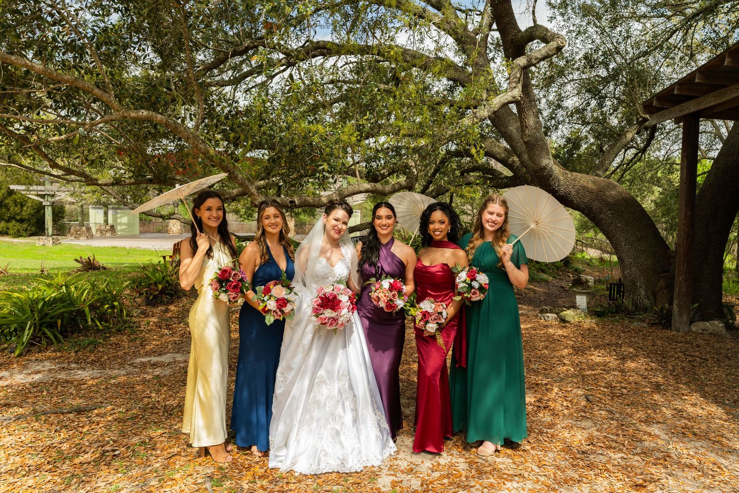Bride in white gown poses with five bridesmaids in colorful dresses holding bouquets under a large oak tree outdoors.
