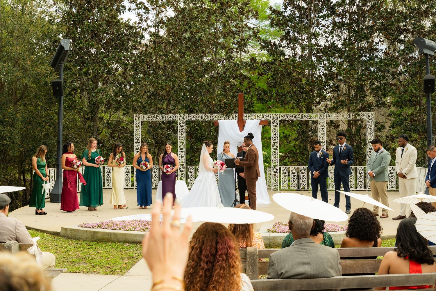 Outdoor wedding ceremony with bride and groom at floral arch, bridesmaids in colorful gowns and groomsmen standing on decorated patio.