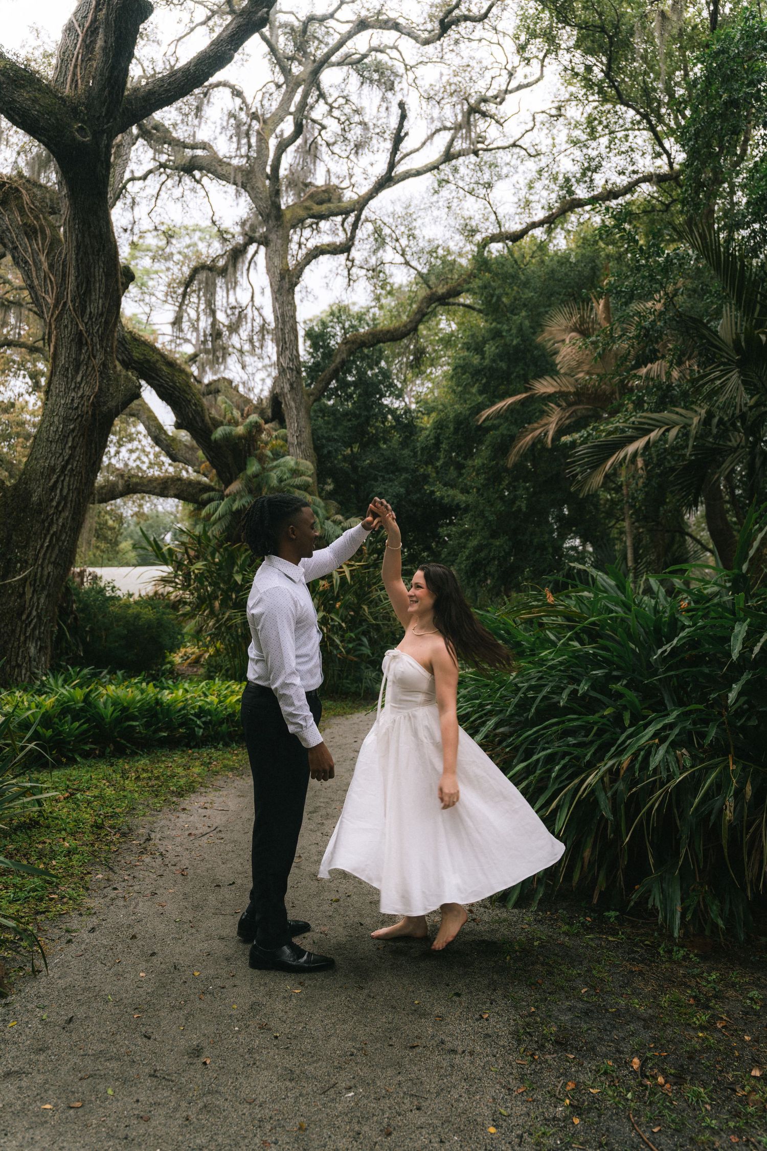 A couple dancing together on a garden path beneath moss-draped oak trees and tropical foliage.