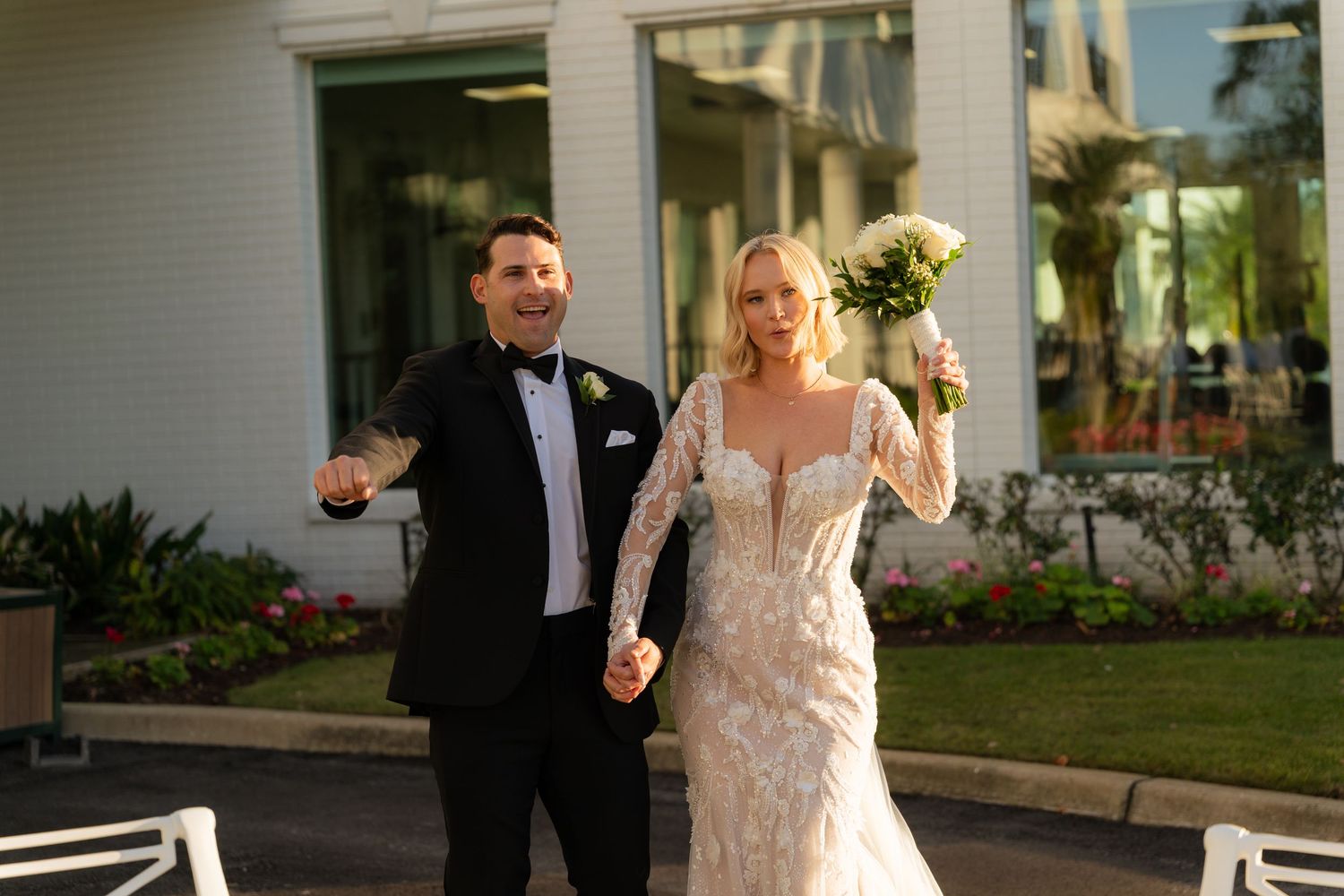 Joyful bride in lace gown holding bouquet and groom in tuxedo celebrating outside after wedding ceremony.