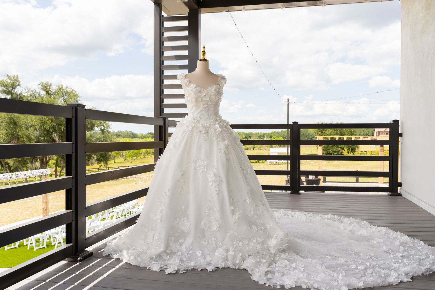 Elegant white ball gown wedding dress with lace appliques displayed on a mannequin on a rustic outdoor balcony.