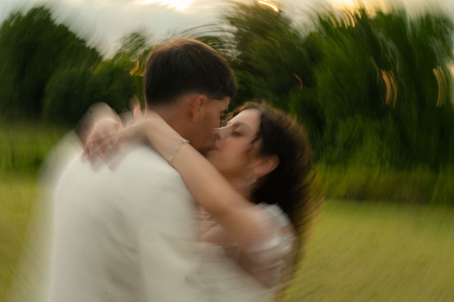 Artistic motion blur photo of newlywed couple sharing a kiss in green outdoor setting.