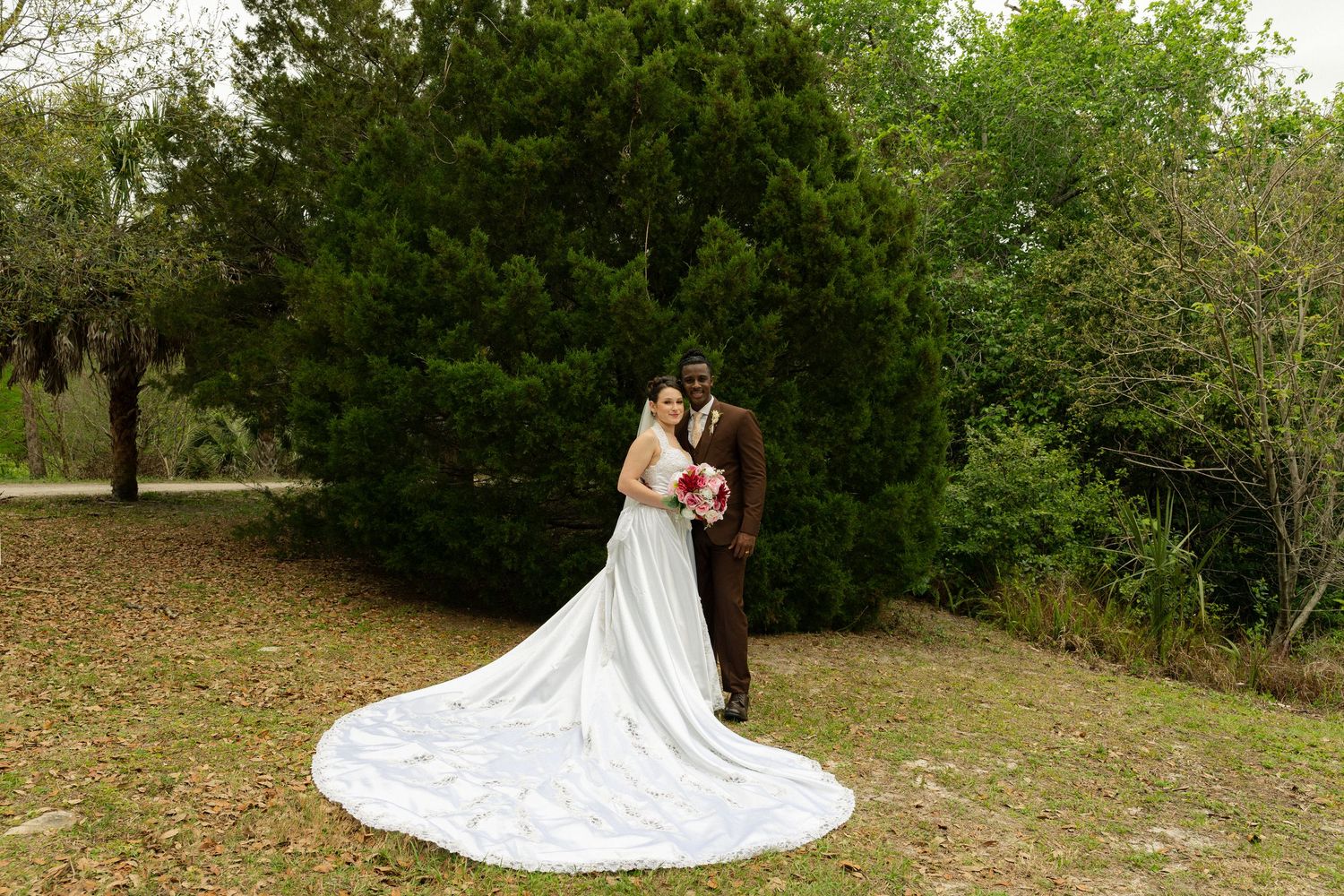 Bride in elegant white gown with long train stands with groom in brown suit outdoors near lush green trees.