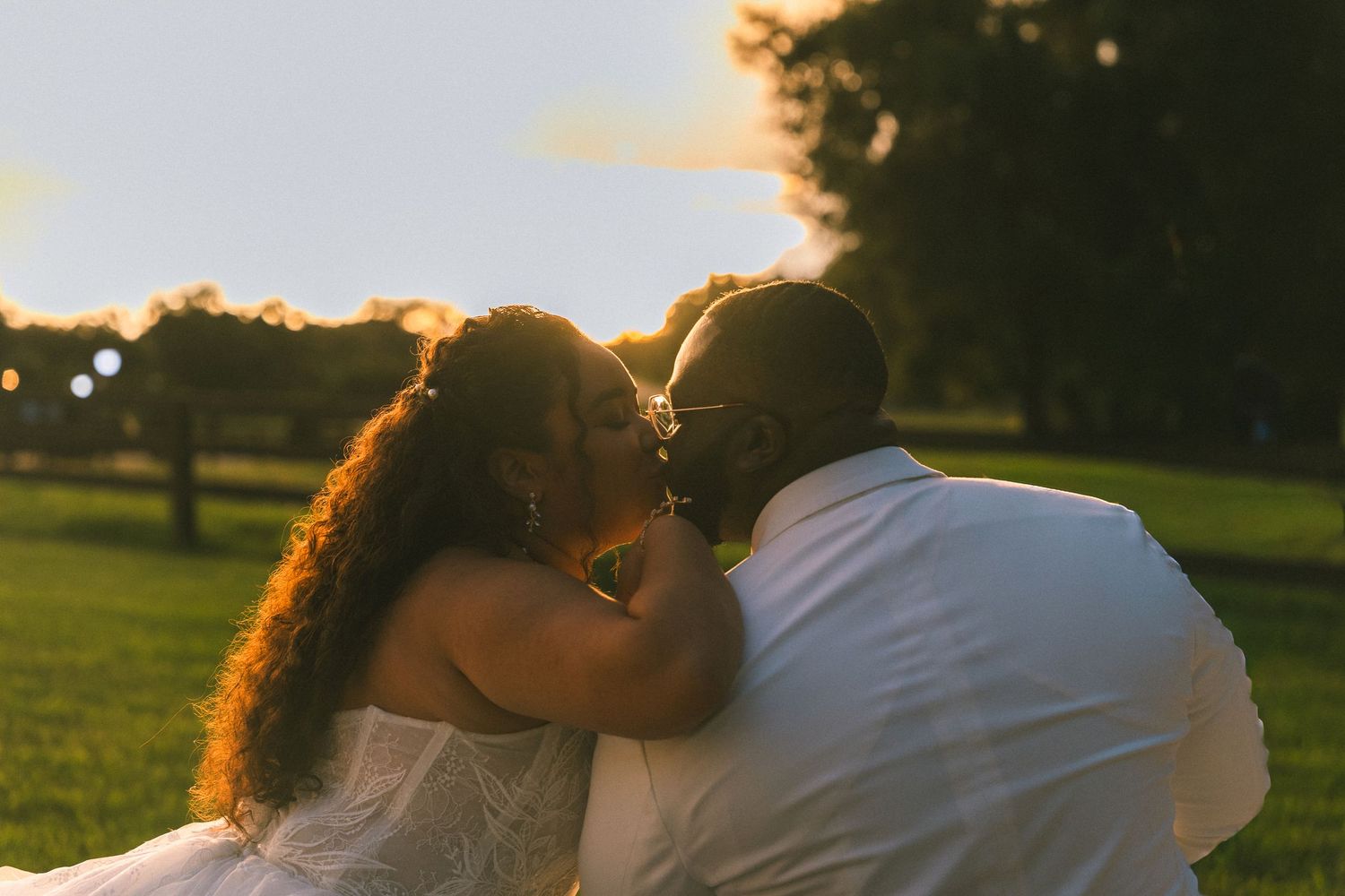 Couple in white outfits sharing a kiss on a grassy lawn at golden hour sunset, with trees and a warm glowing sky behind them.