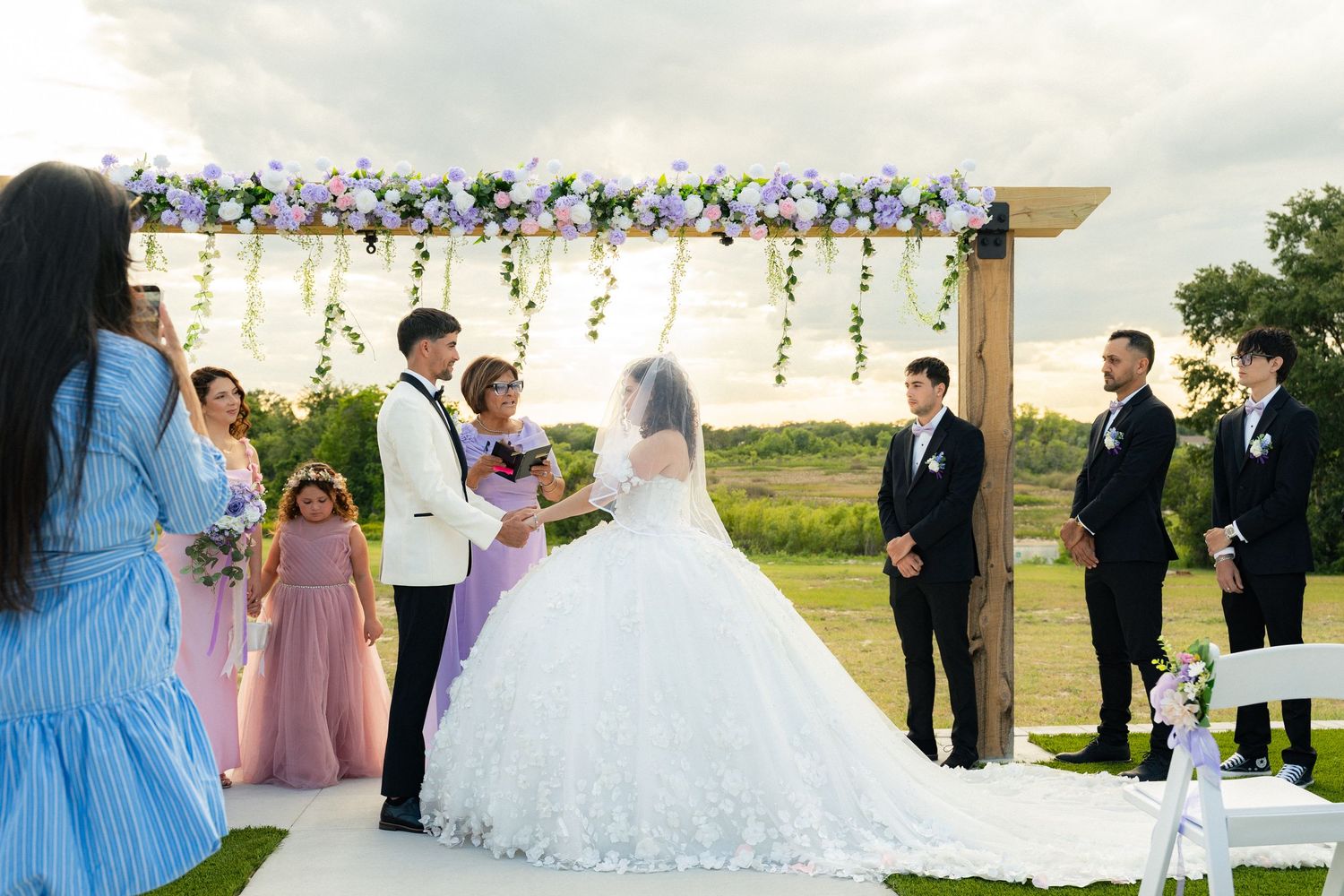 Couple exchanges vows under floral arch at outdoor wedding ceremony with bridal party at golden hour.