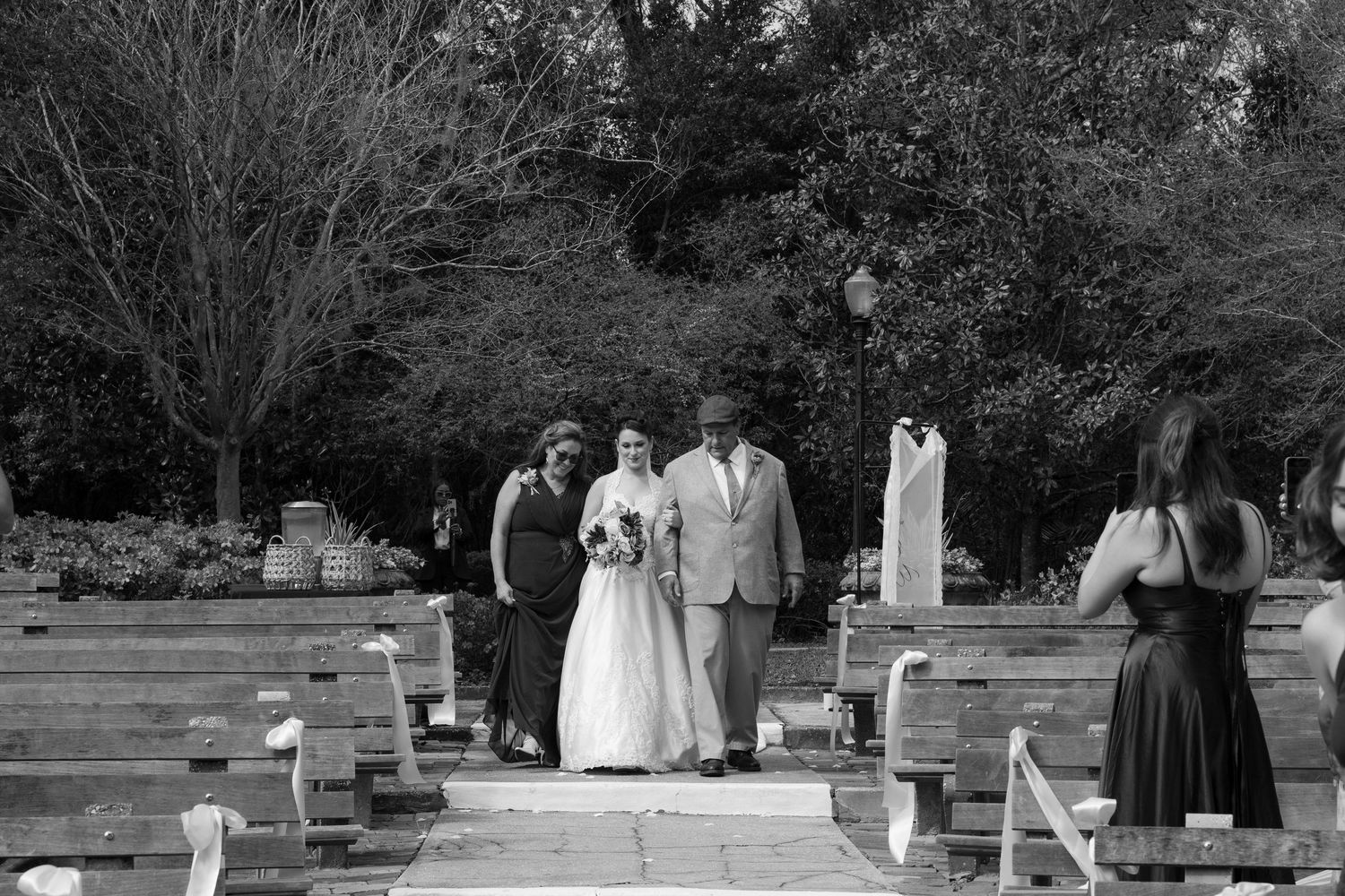 Black and white photo of a bride walking down the aisle with family at an outdoor wedding ceremony.