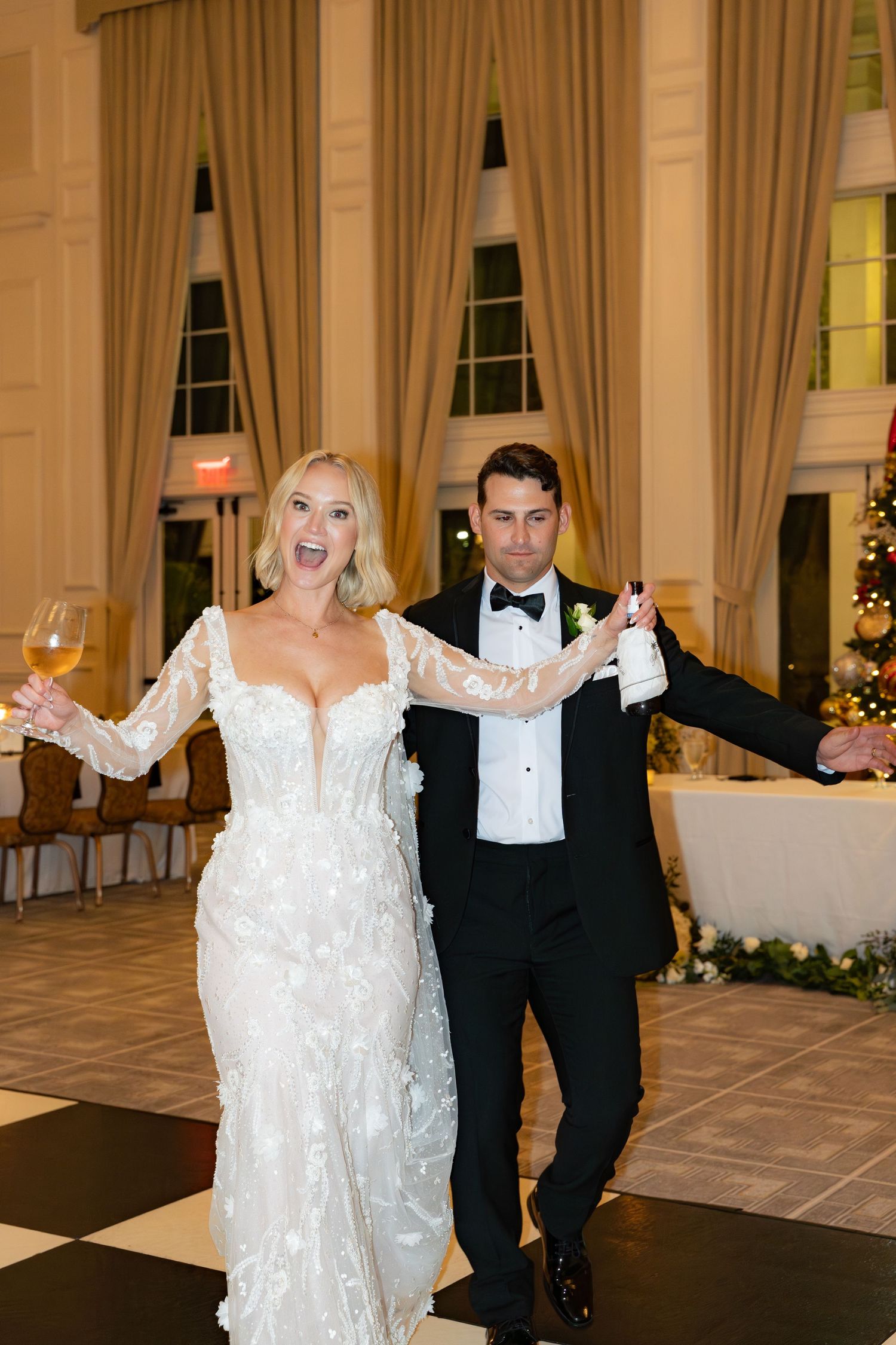Bride and groom make joyful grand entrance at wedding reception, bride holding wine glass in lace gown, groom in black tuxedo.