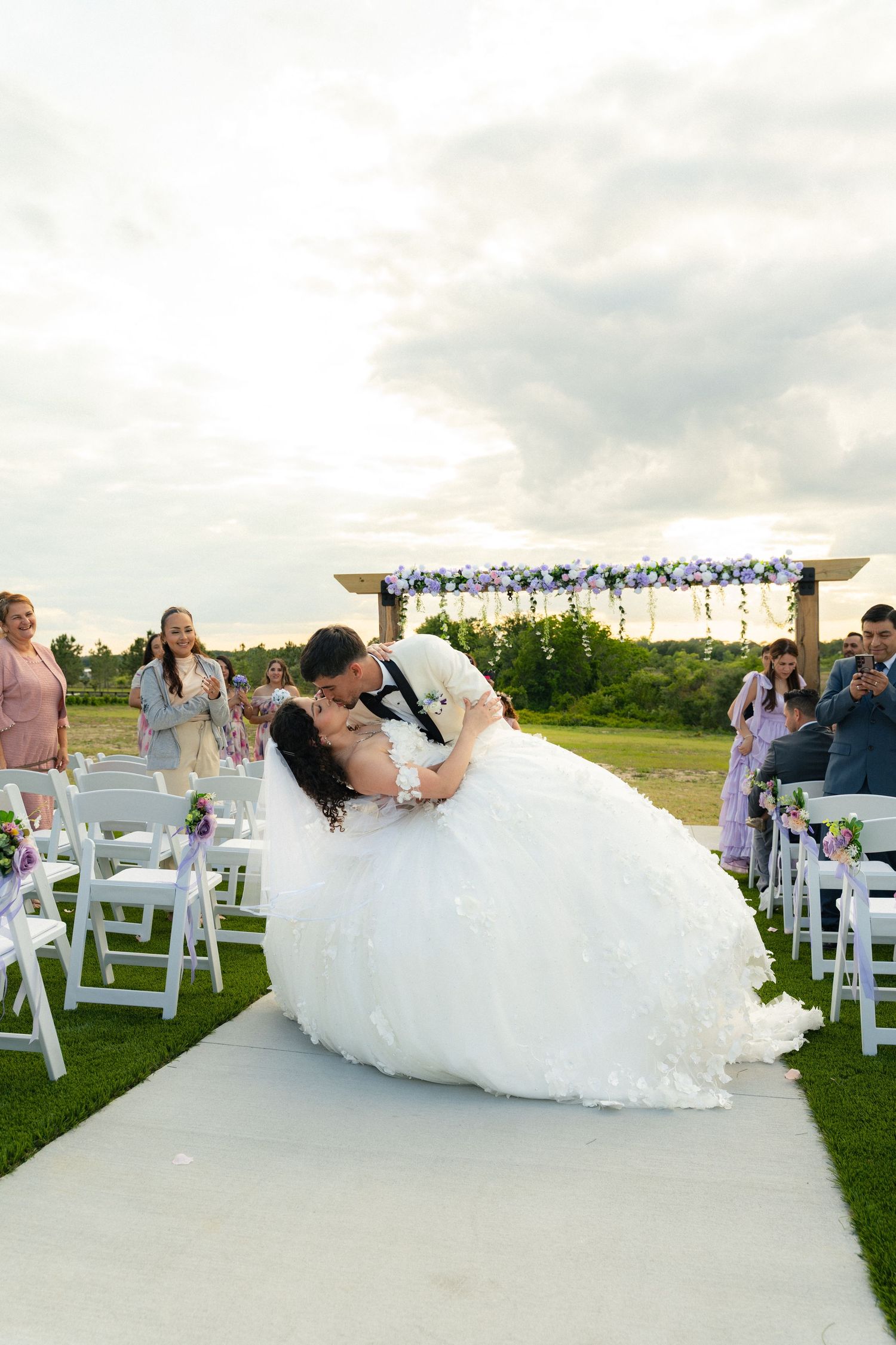 Groom dips bride for a kiss at outdoor wedding ceremony with guests and floral arch behind.