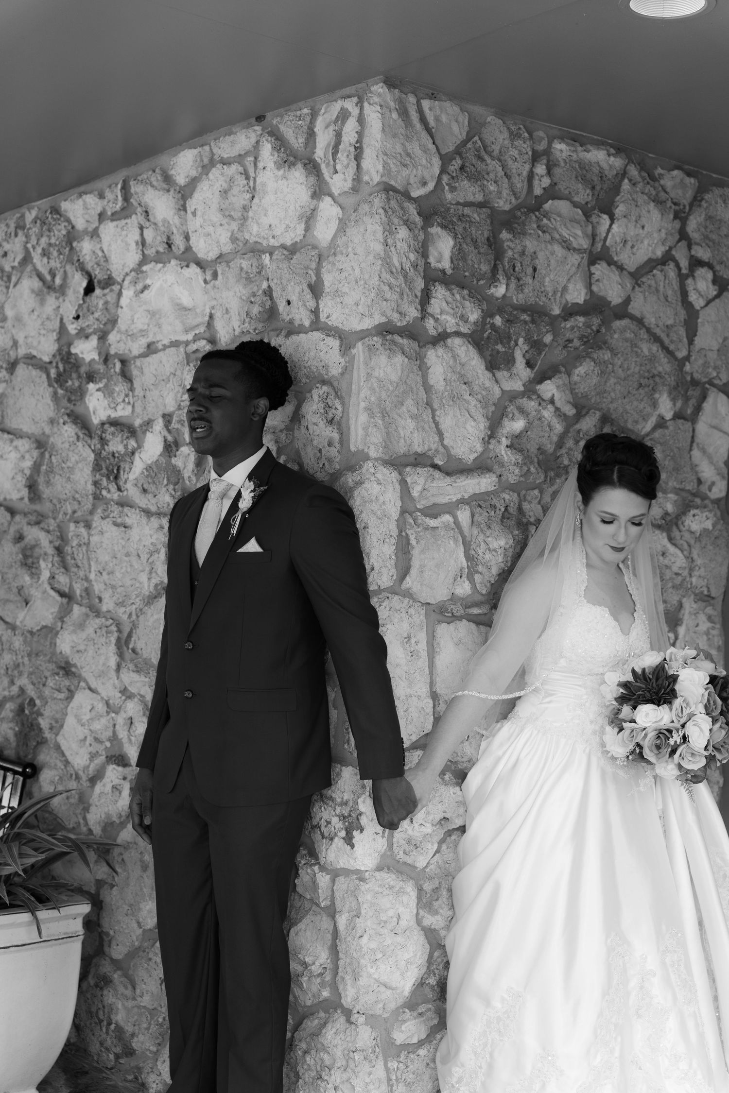 Bride and groom hold hands in black and white wedding portrait against rustic stone wall.