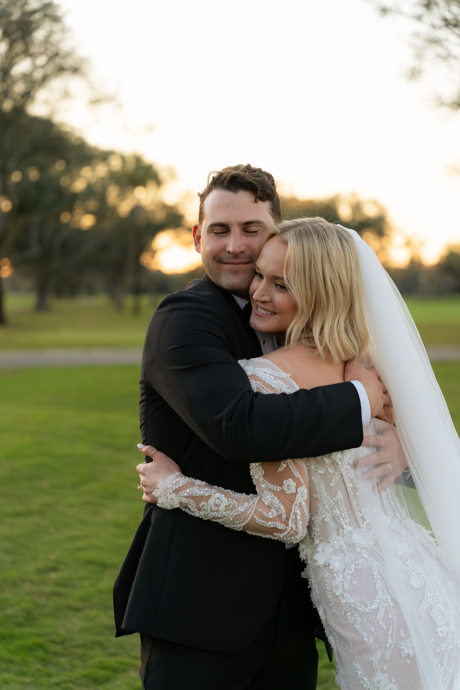 A newlywed couple shares a tender embrace at golden hour on a lush green lawn after their wedding.