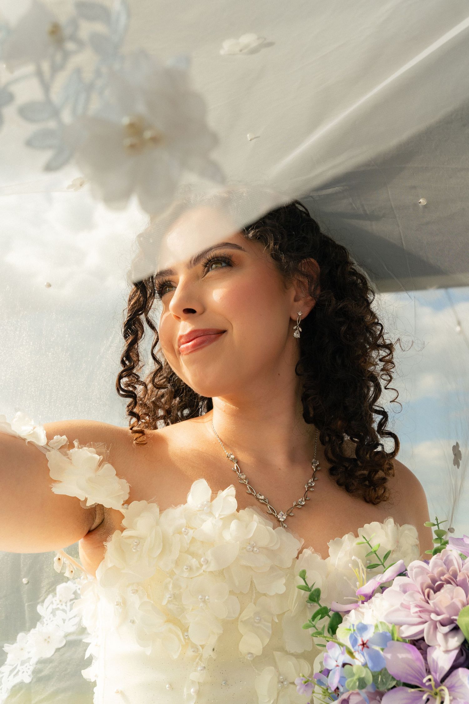 Beautiful curly-haired bride in strapless floral gown holds purple bouquet beneath flowing white veil outdoors.