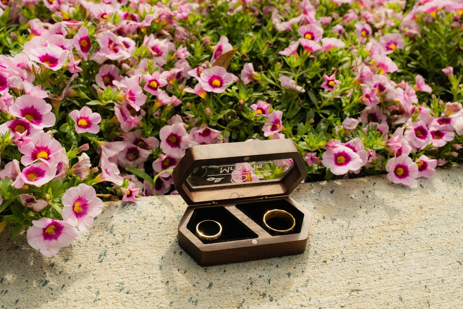 Two gold wedding bands in an open wooden ring box displayed on stone surface with pink petunia flowers in background.
