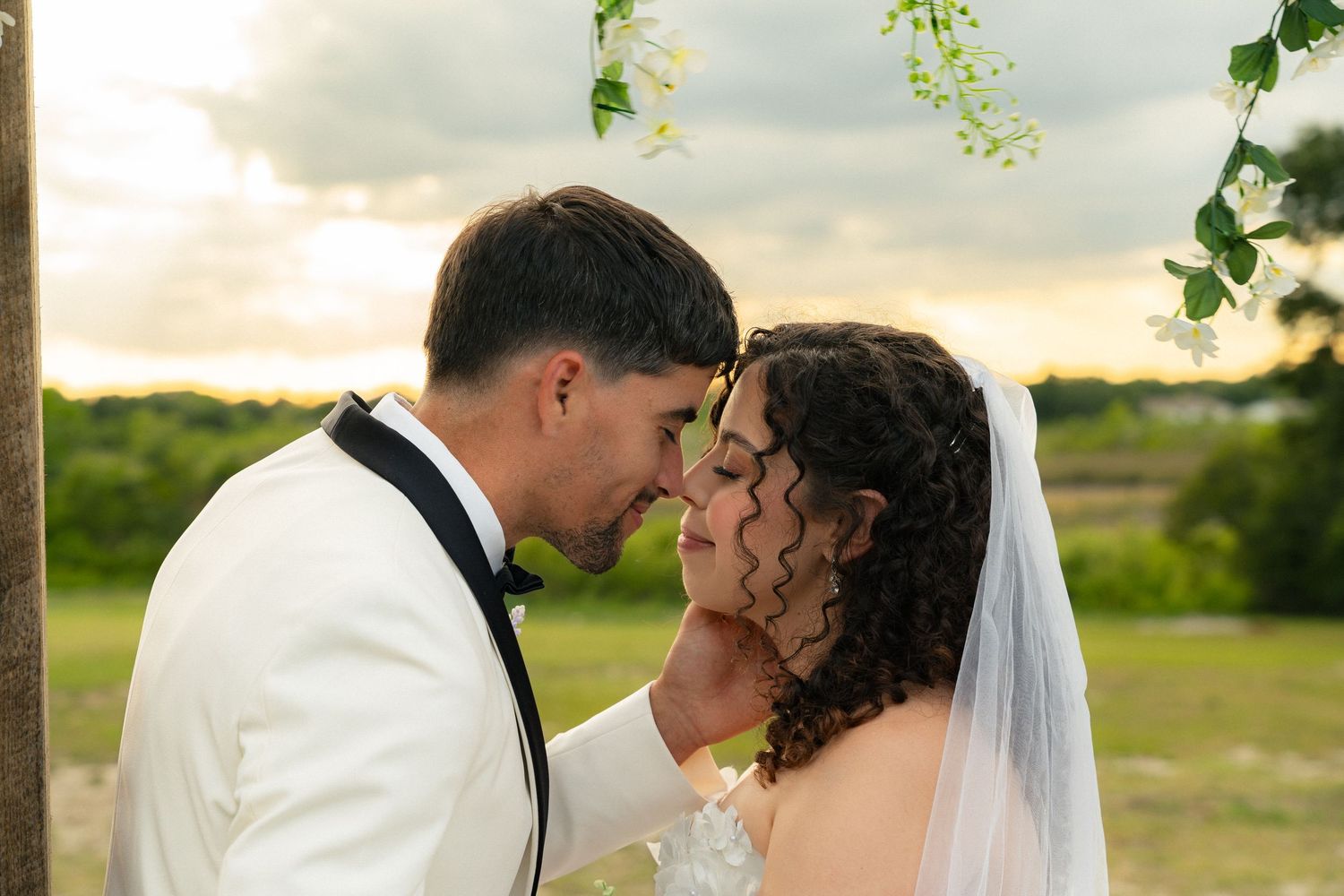 Bride and groom share an intimate forehead touch moment under a floral arch at sunset outdoor ceremony.