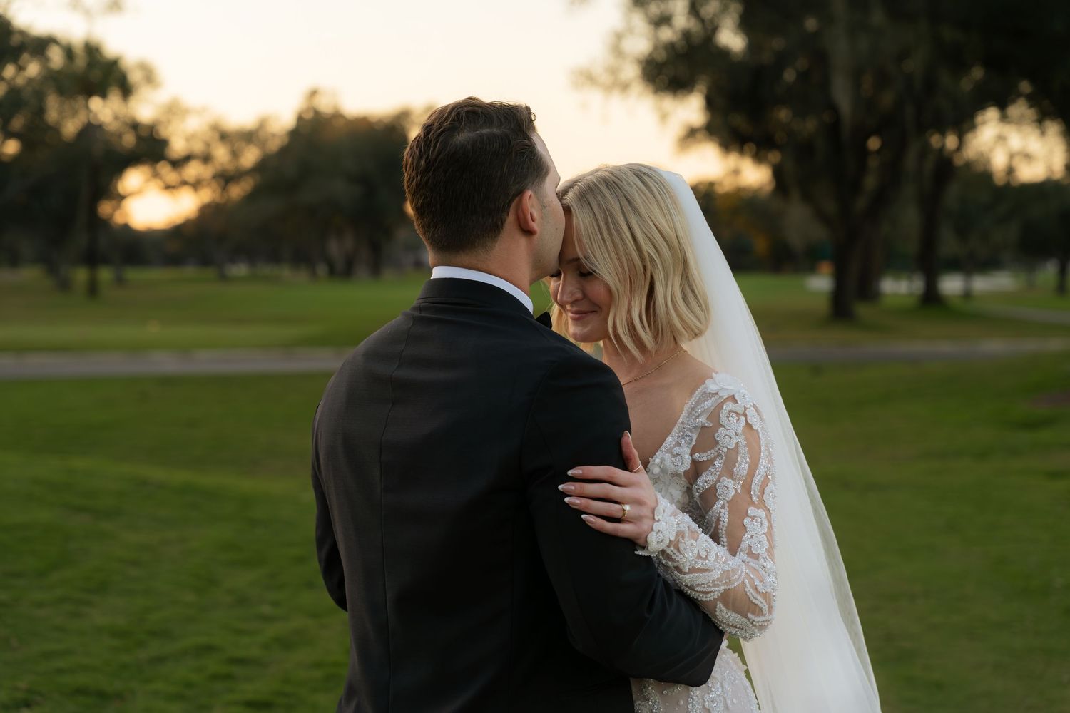 Bride and groom share romantic embrace at sunset on green golf course after wedding ceremony.
