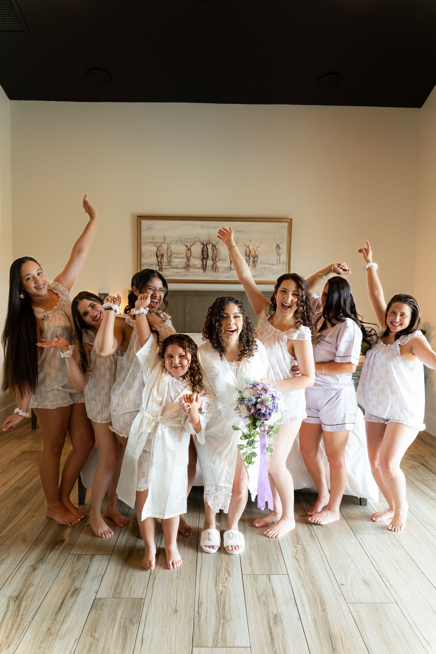 Bride in white robe holds purple bouquet surrounded by cheerful bridesmaids in pajamas celebrating before the wedding.