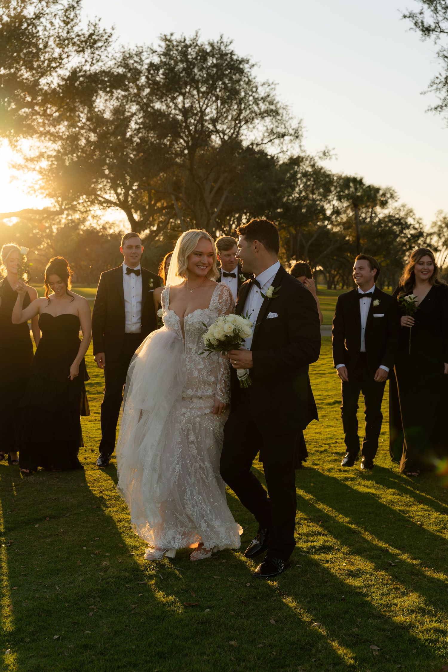 Bride and groom walk together at golden hour sunset wedding with bridal party dressed in black formal attire.