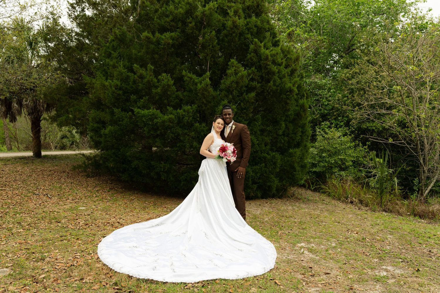Bride in a long white gown with train and groom posing before tall green trees.