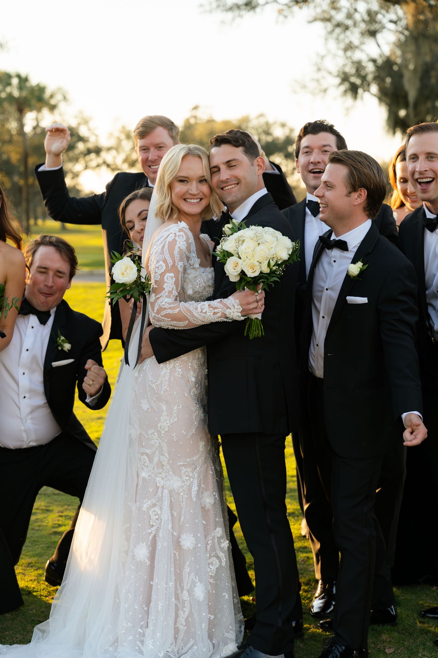 A joyful bride and groom celebrate with their wedding party on a sunny green lawn after the ceremony.