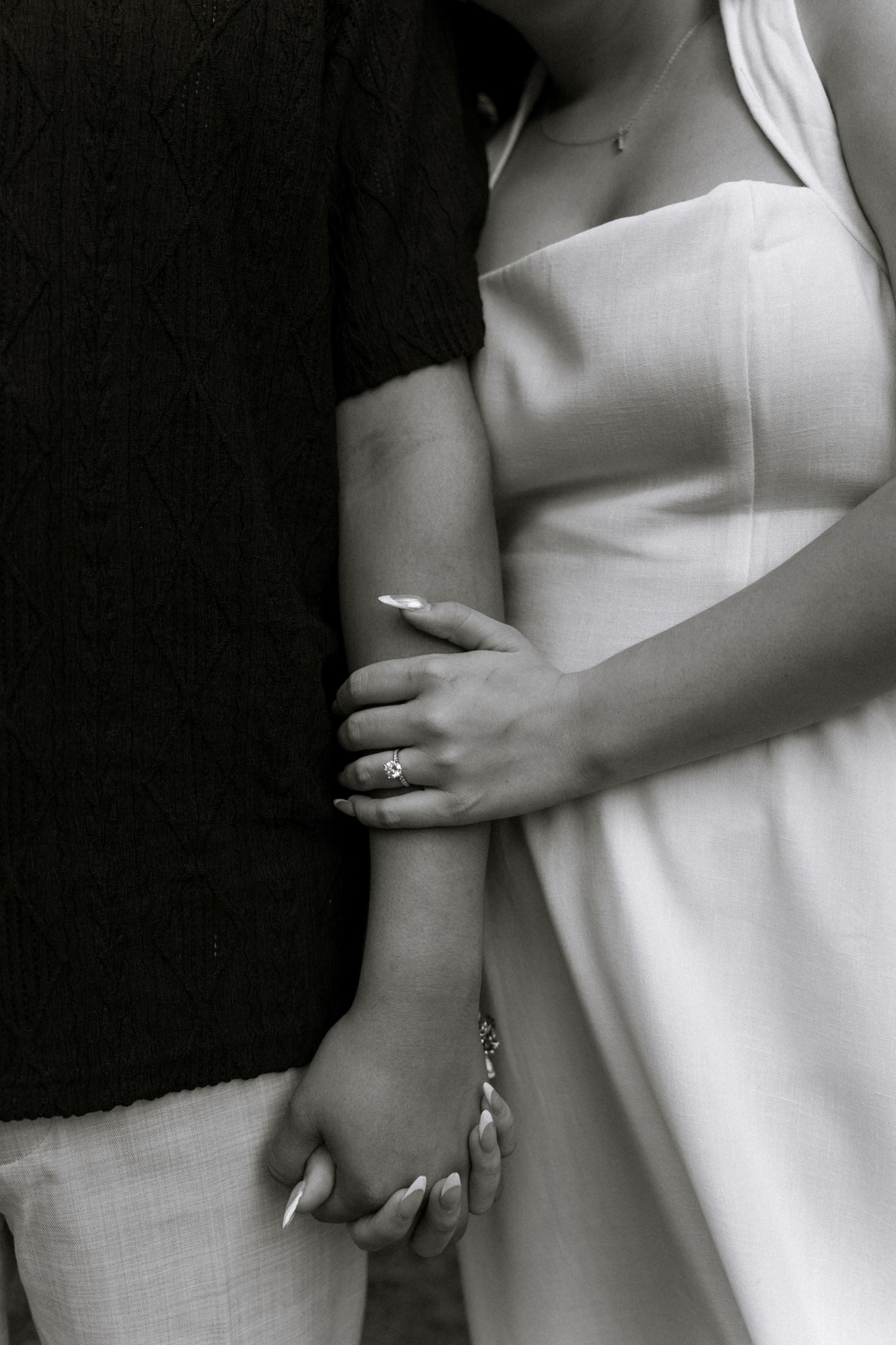 Black and white photo of bride and groom holding hands, showing wedding ring and white gown.