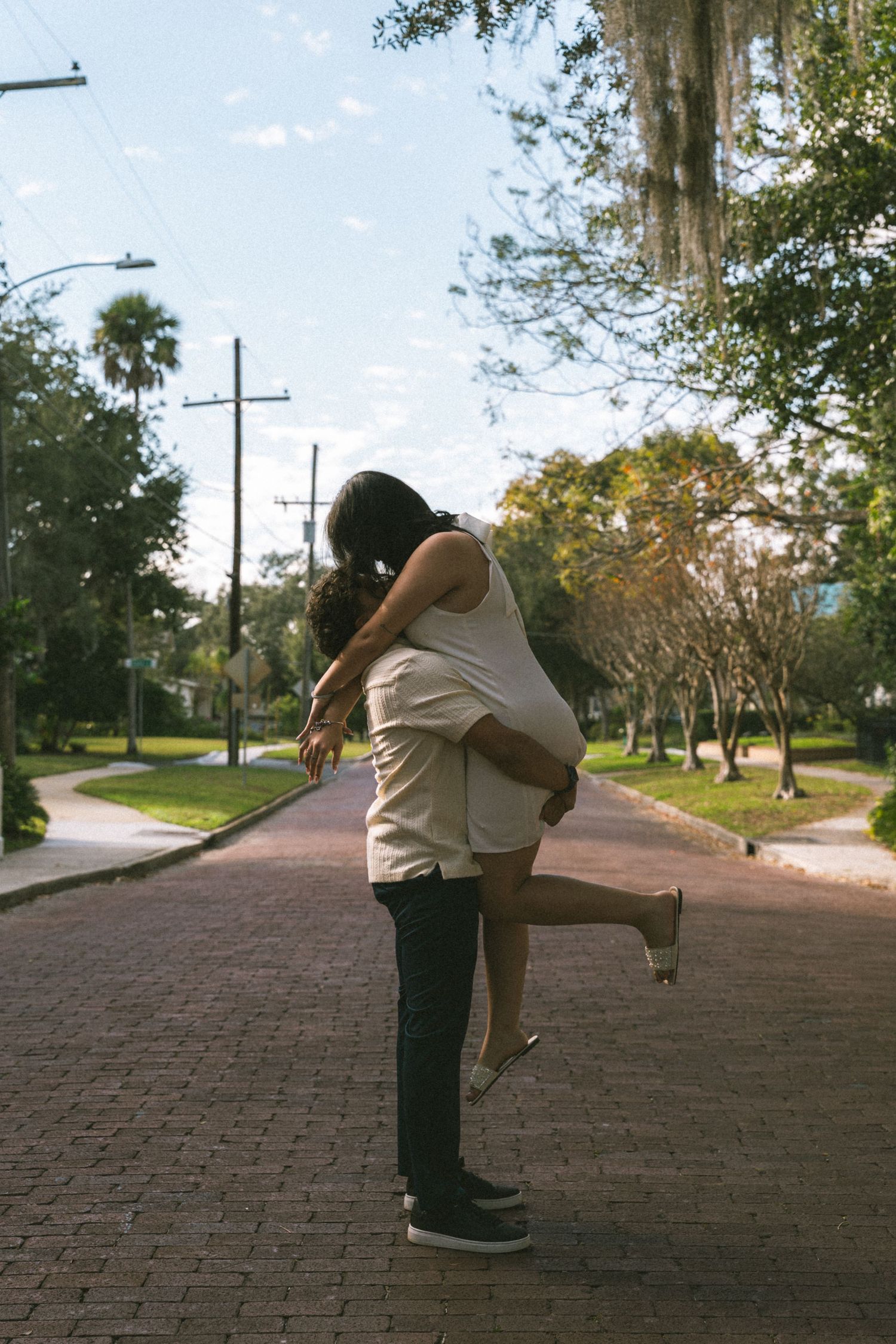 A couple embraces in a tender hug while standing on a brick pathway lined with trees and utility poles.