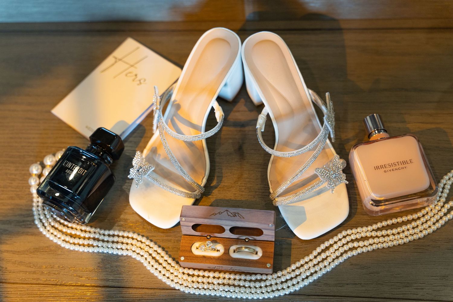 White strappy heeled sandals with crystal bow, pearl necklace, perfume, and jewelry box on wooden surface.