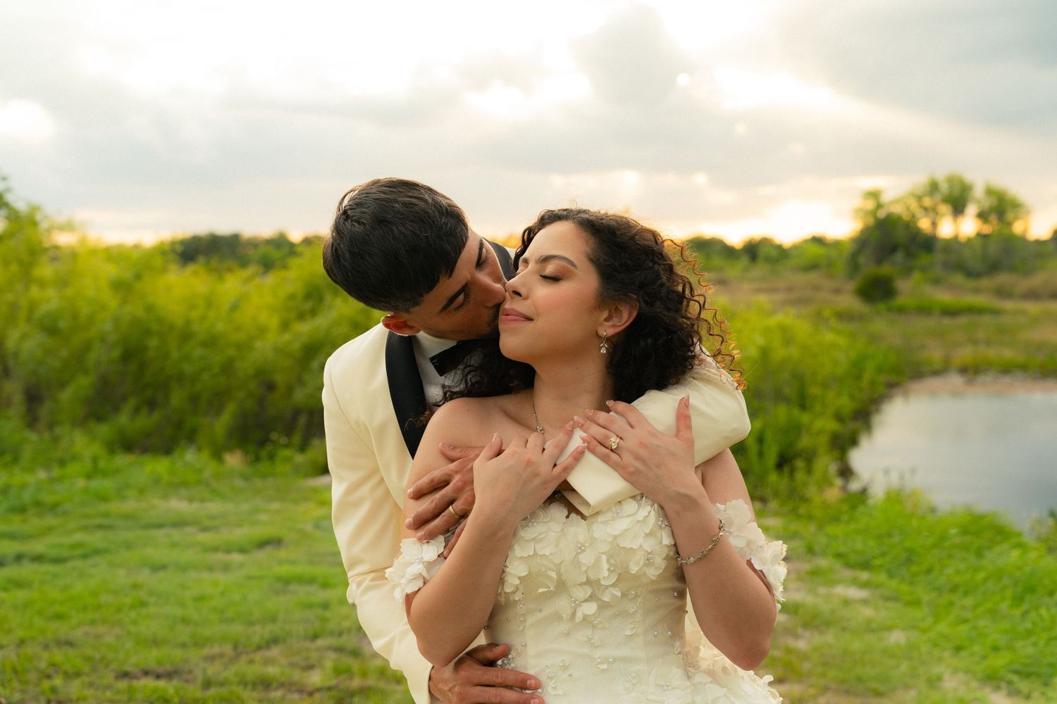 Groom tenderly embracing bride from behind in a lush green outdoor setting near a serene pond.
