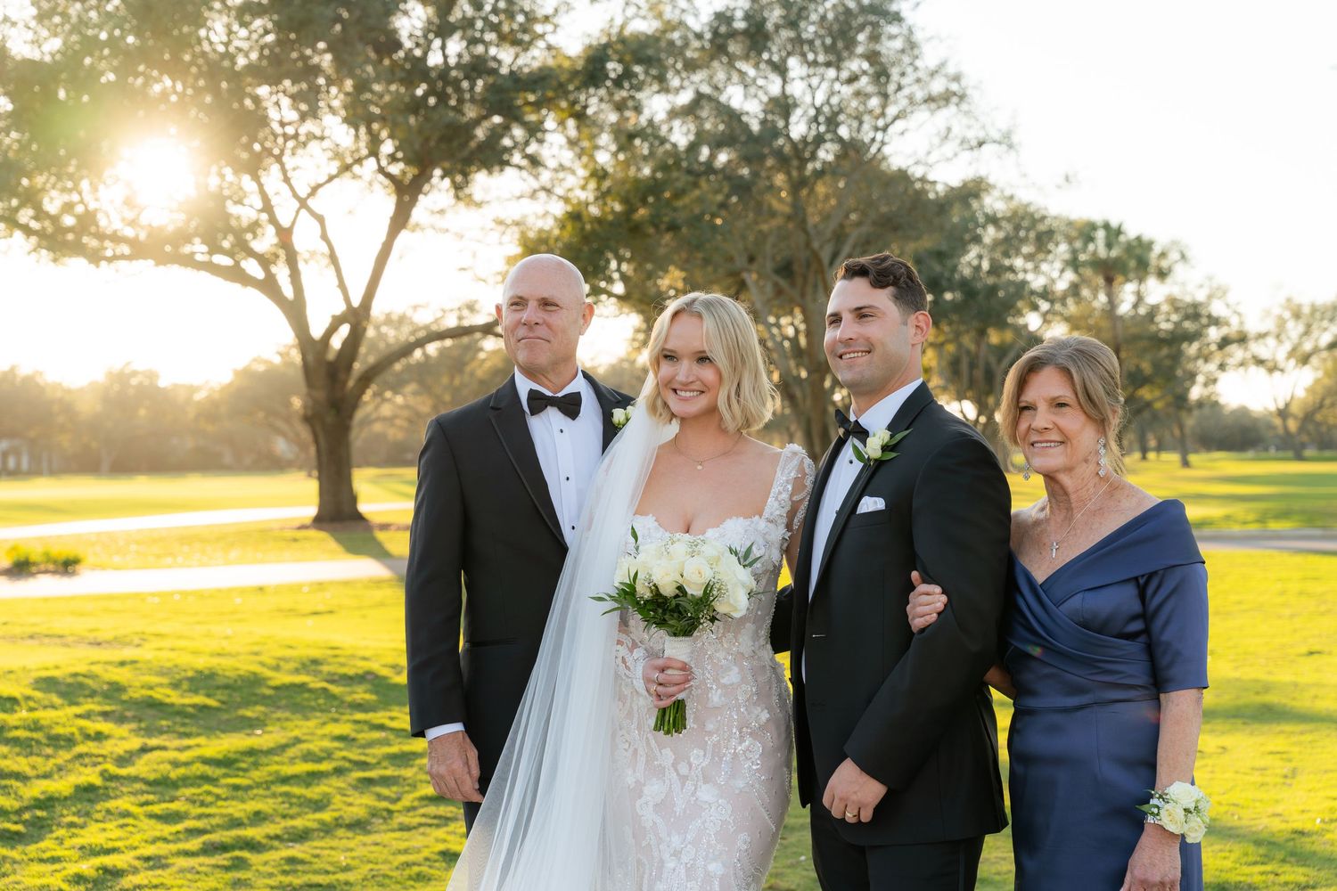 Bride and groom pose with parents at golden hour on a lush green golf course.
