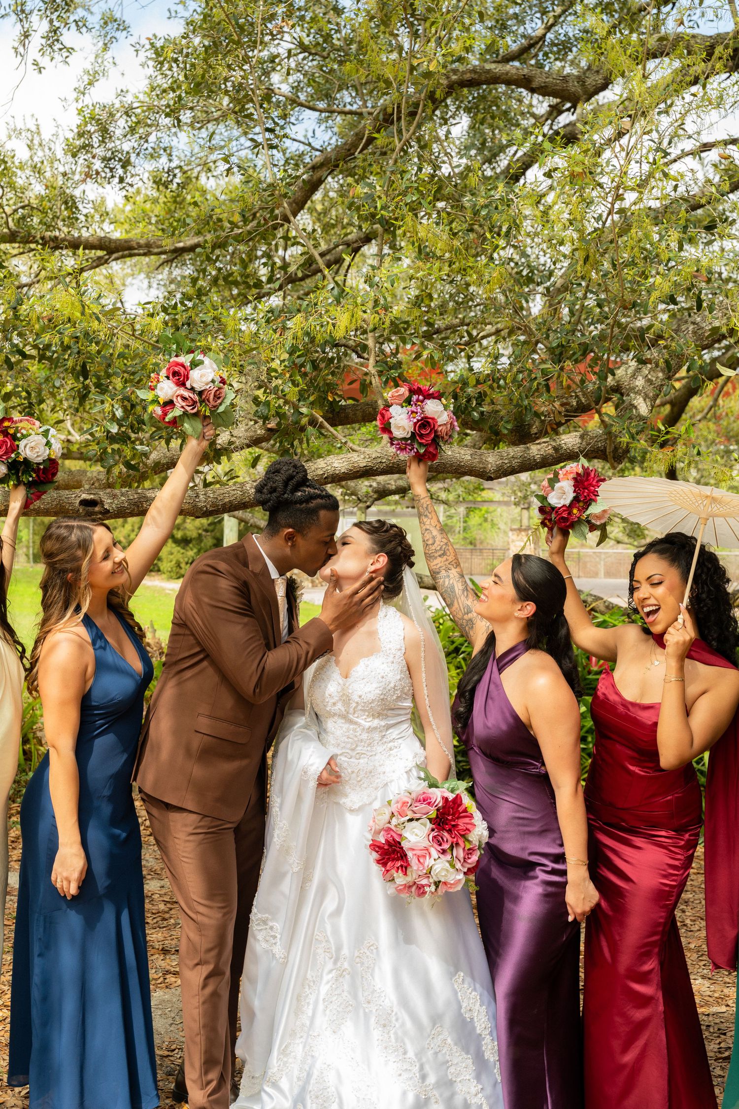 Bride and groom share a kiss as bridesmaids in jewel-toned gowns celebrate and raise bouquets under oak tree.